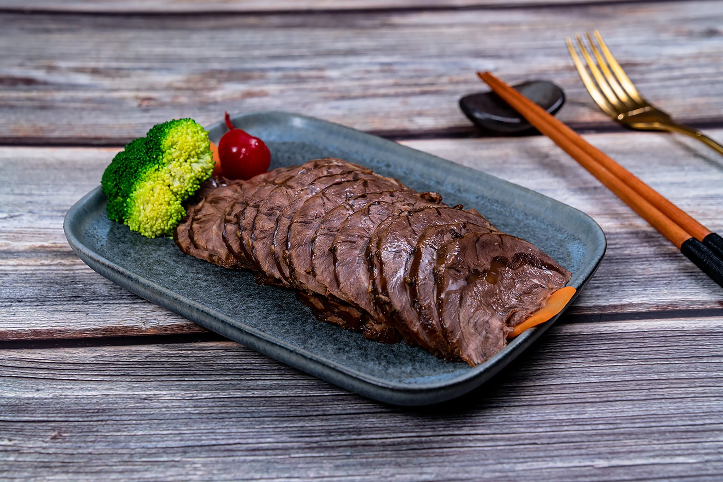 Slices of cooked beef sirloin on a gray plate with broccoli and a cherry tomato, set on a wooden table with chopsticks and a spoon in the background.