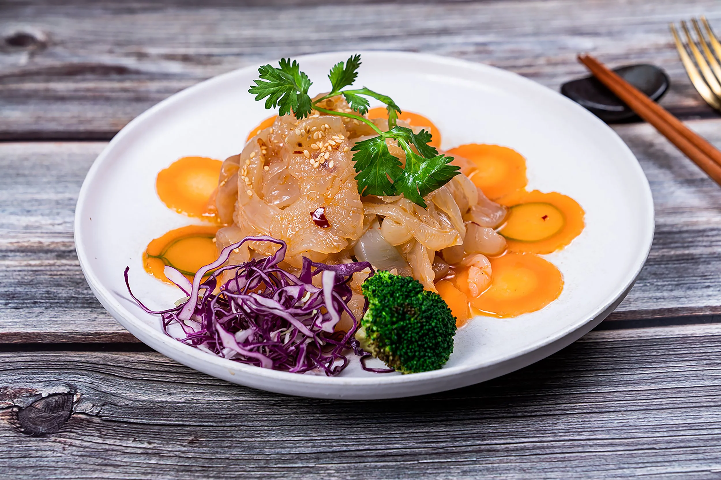 A white plate with a serving of sashimi garnished with fresh cilantro, sesame seeds, and purple shredded cabbage, accompanied by slices of carrot, broccoli, and cucumber, on a rustic wooden table.