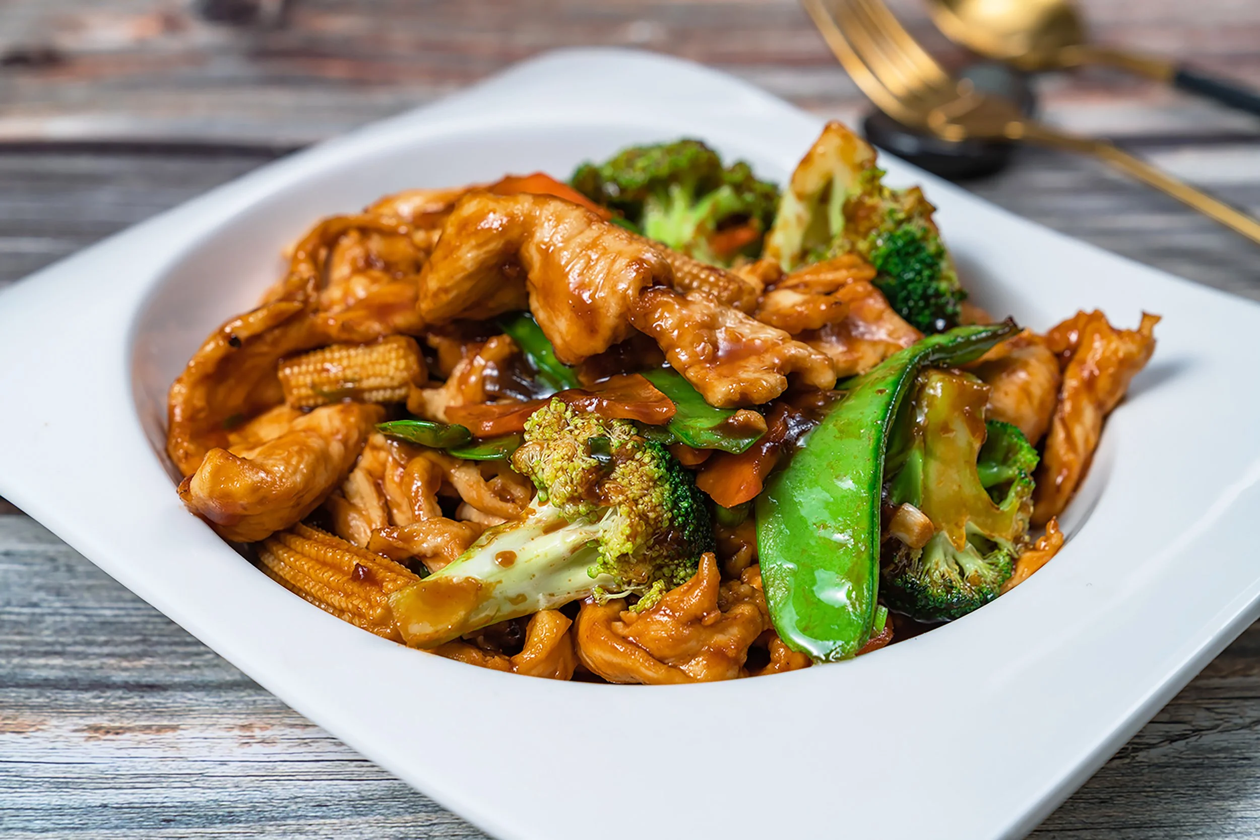 A white bowl of stir-fried vegetables and meat with broccoli, carrots, snow peas, and baby corn, covered in a savory brown sauce.