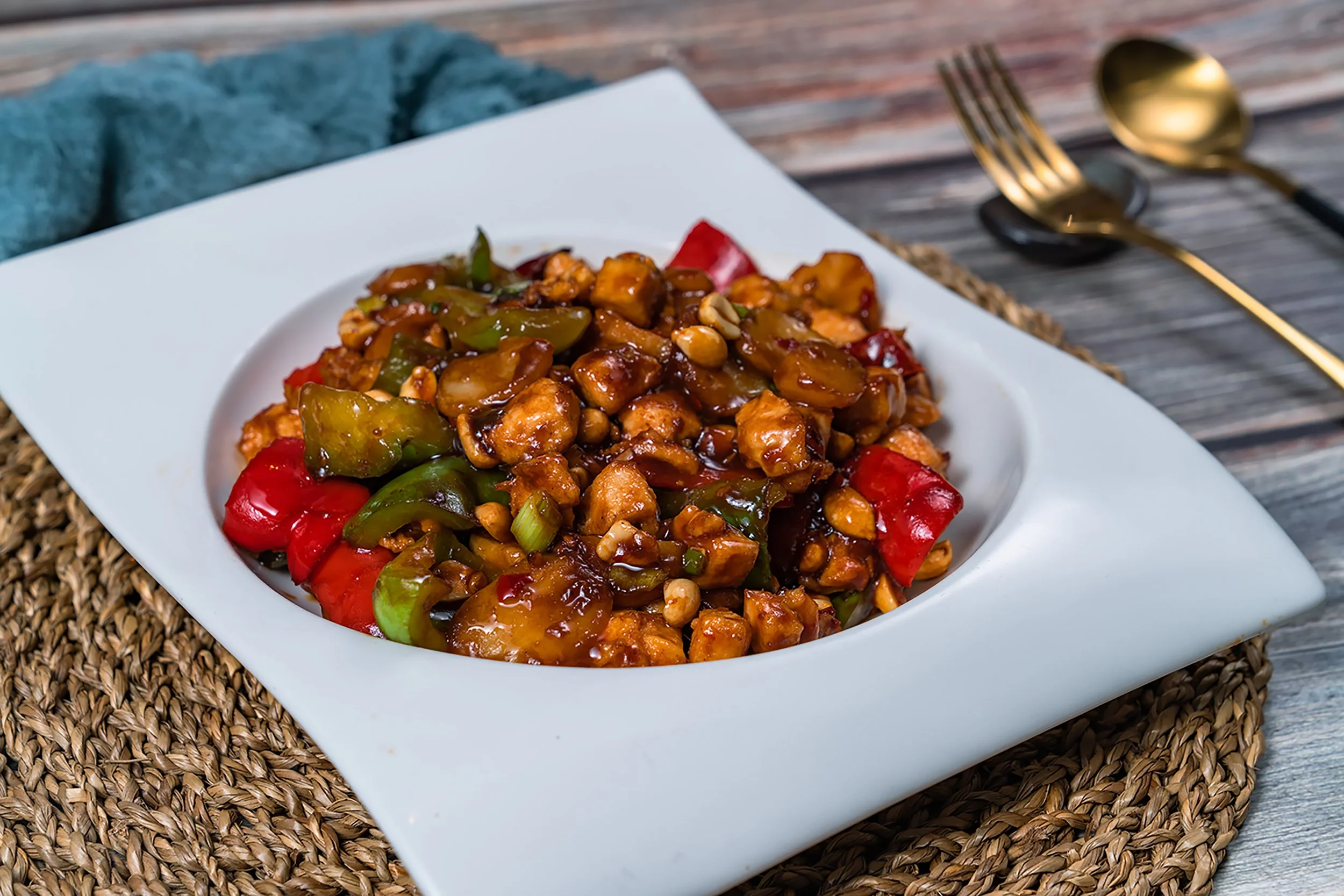 A plate of sweet and sour chicken with vegetables on a white dish, placed on a woven placemat on a rustic wooden table, with a gold spoon and fork to the side.