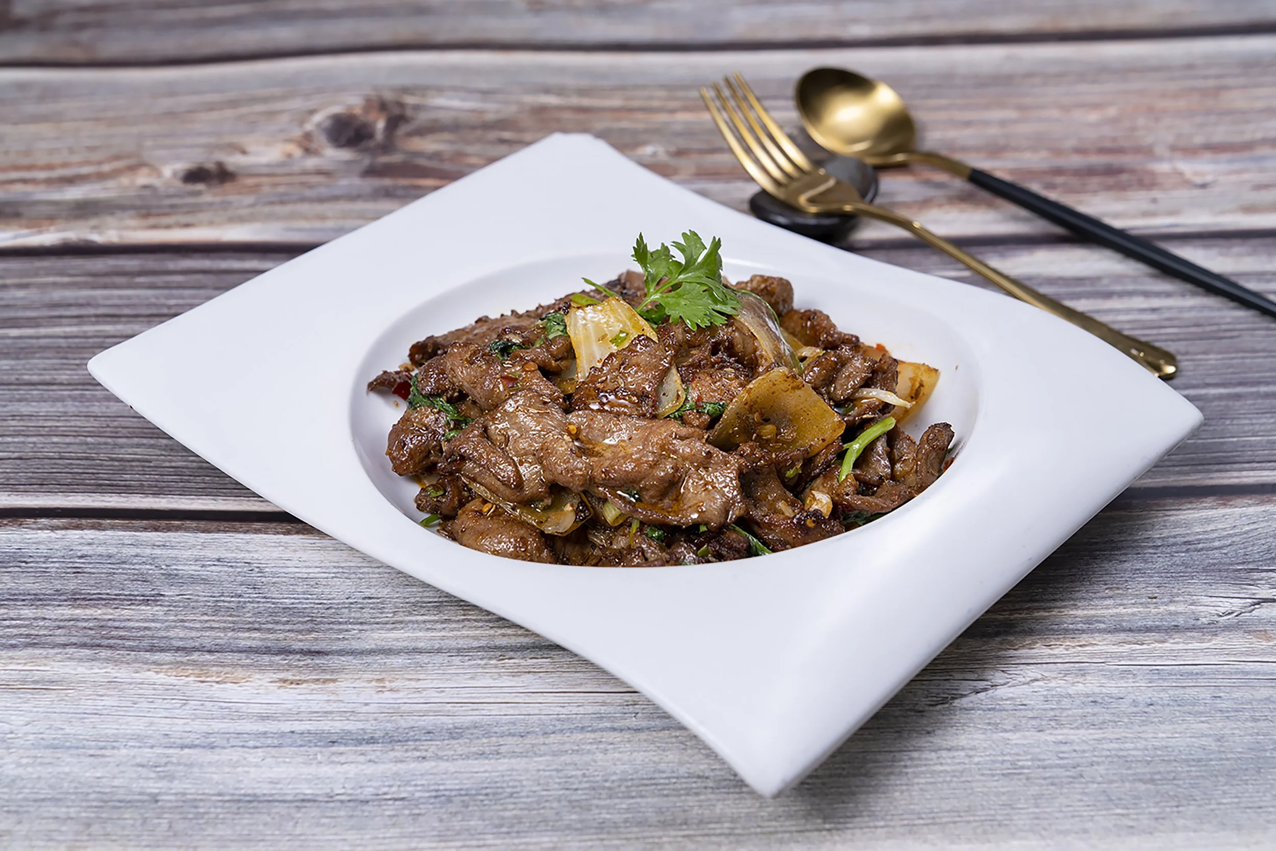 A white rectangular plate with a serving of stir-fried beef and vegetables garnished with cilantro, placed on a wooden table with a gold spoon and fork beside it.