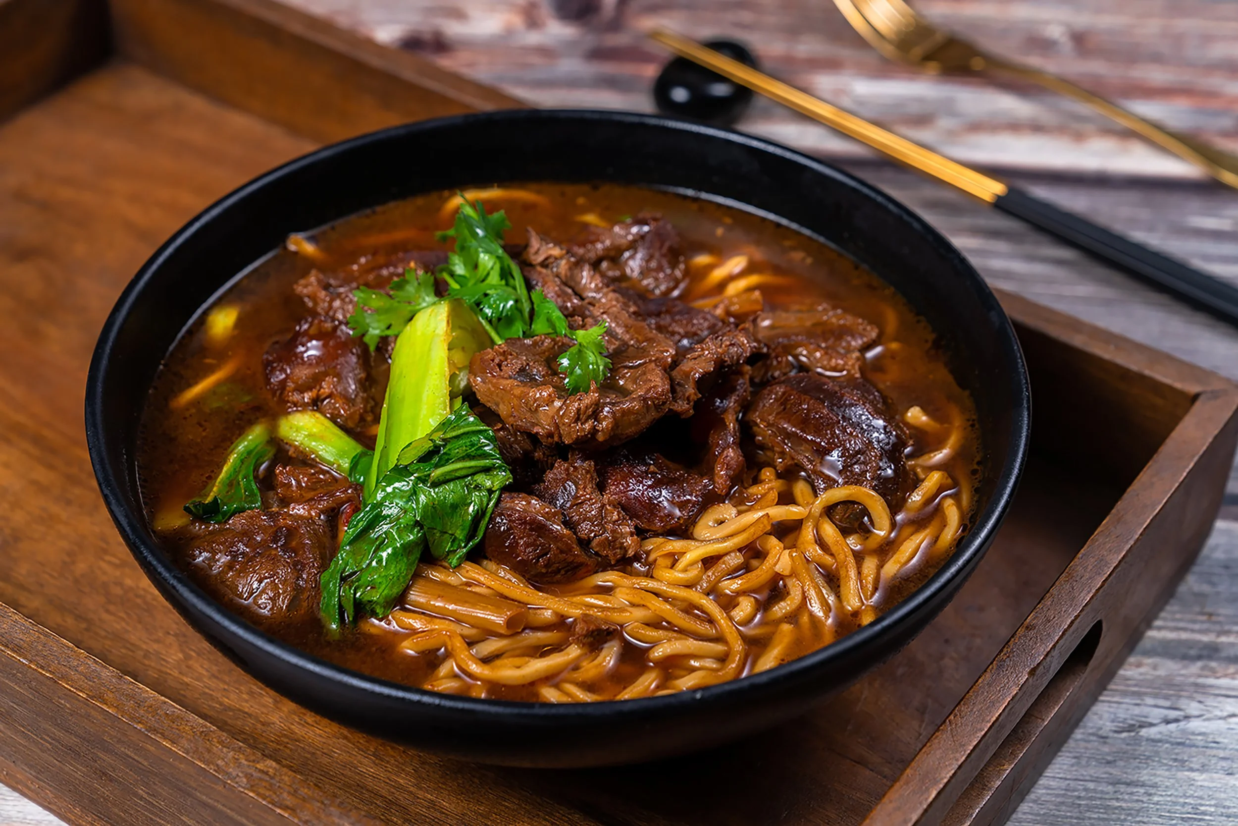 A black bowl of Asian noodle soup with beef, green vegetables, and cilantro, placed on a wooden tray on a rustic wooden table.