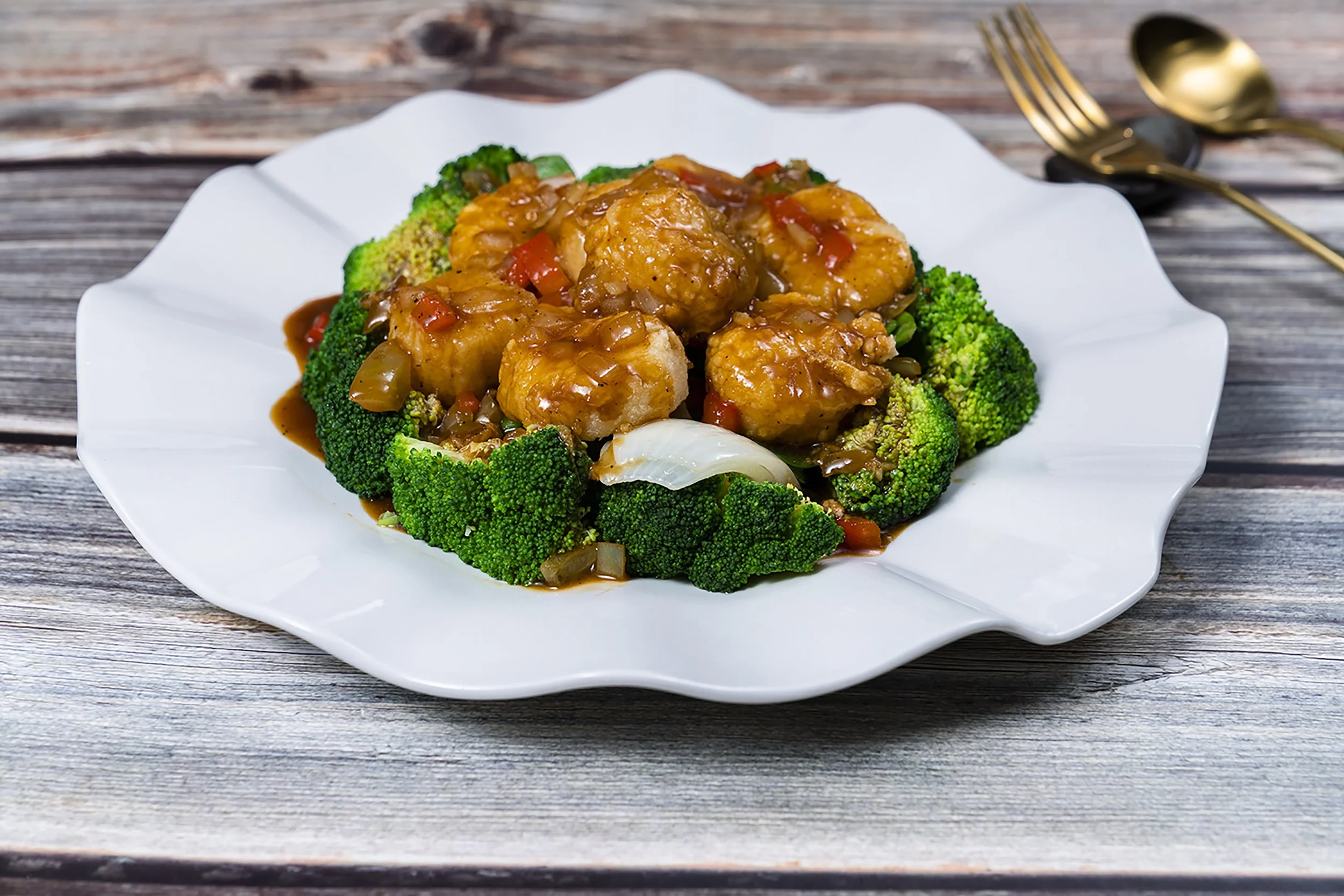 Dish of broccoli florets topped with saucy chicken pieces on a white plate, with a gold fork and spoon placed on a wooden surface in the background.