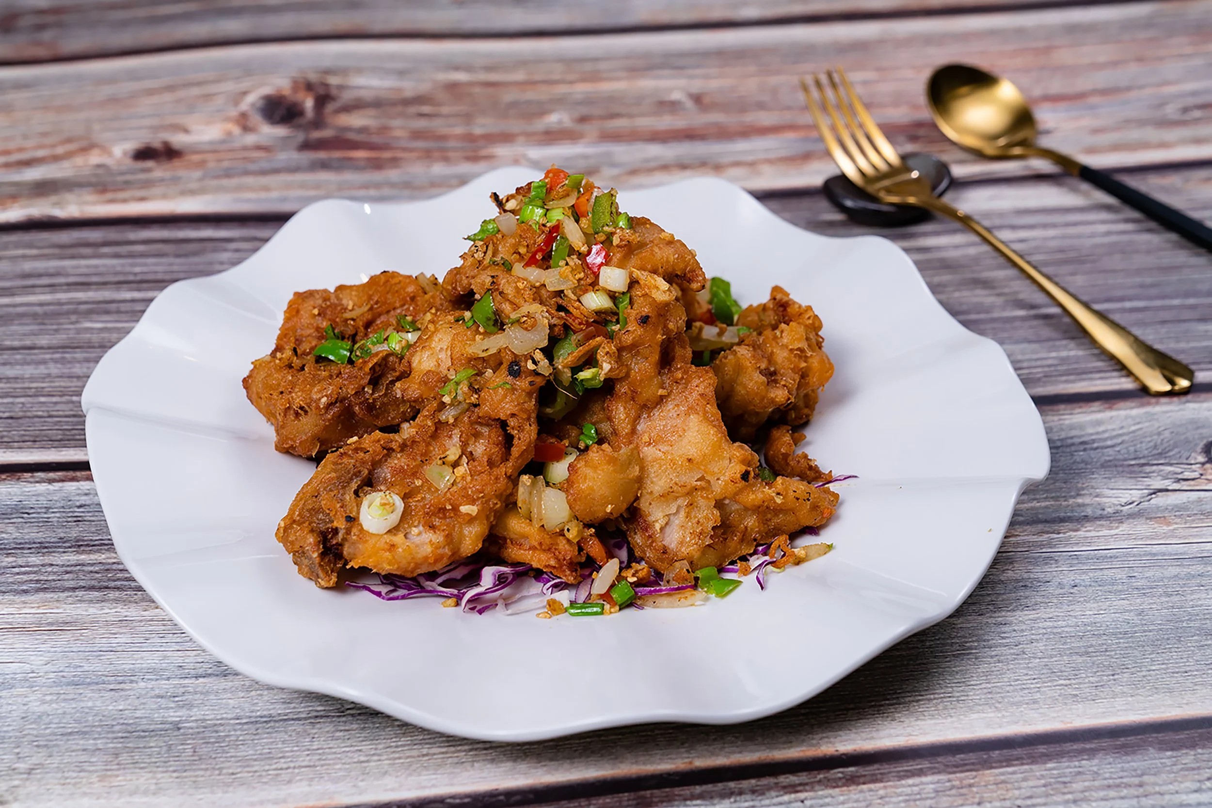 Fried chicken topped with chopped green onions and red chili peppers on a white plate, placed on a wooden surface with gold fork and spoon in the background.