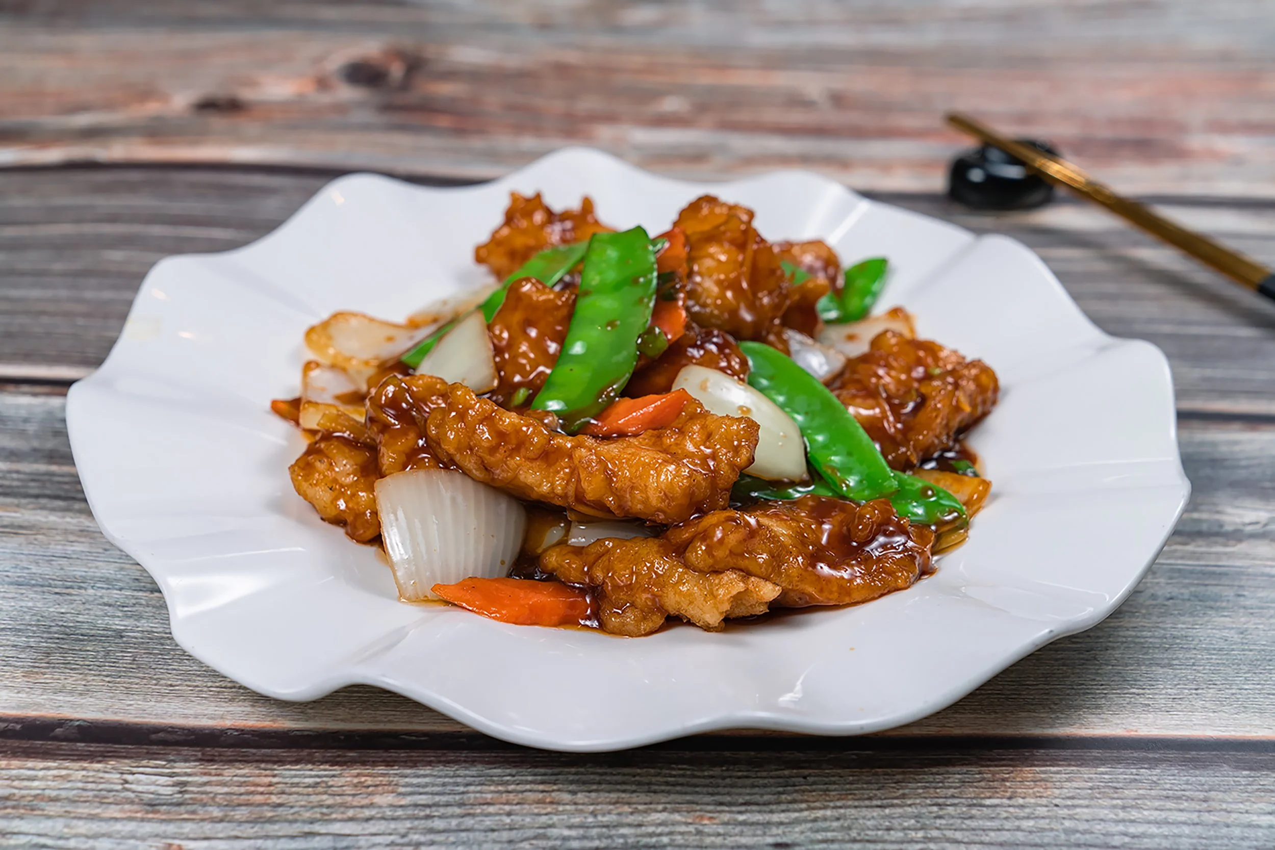A white decorative plate with sweet and sour chicken garnished with green onions and vegetables, placed on a wooden surface, with chopsticks on the side.