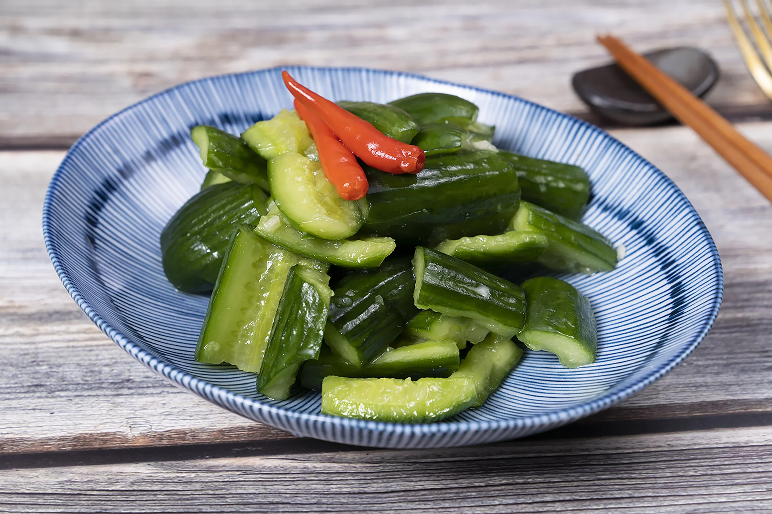 A blue and white bowl filled with chopped cucumbers and two red chili peppers on top, placed on a wooden table.