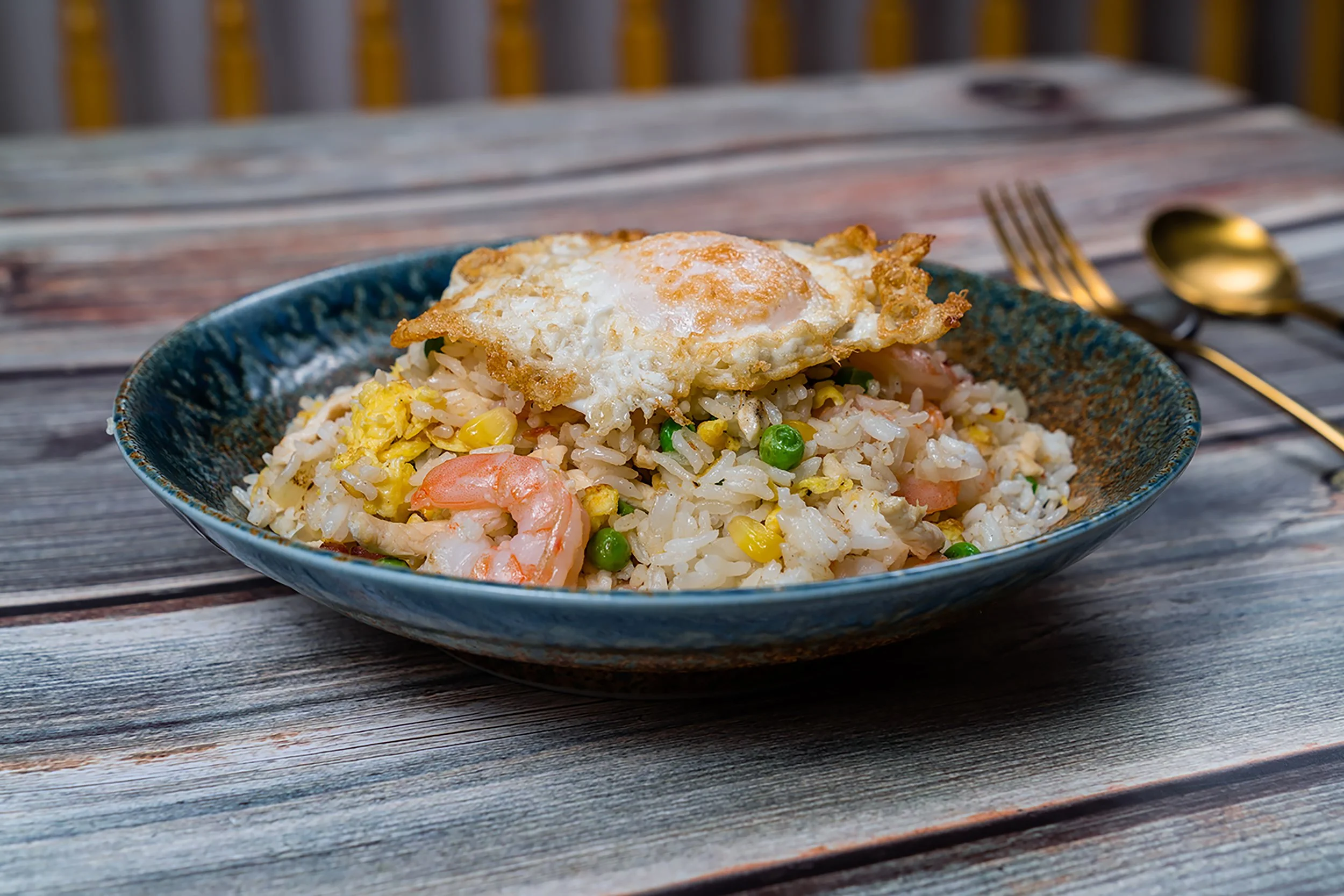 A bowl of fried rice with shrimp, egg, peas, and corn, topped with a fried egg, on a wooden table with gold utensils in the background.
