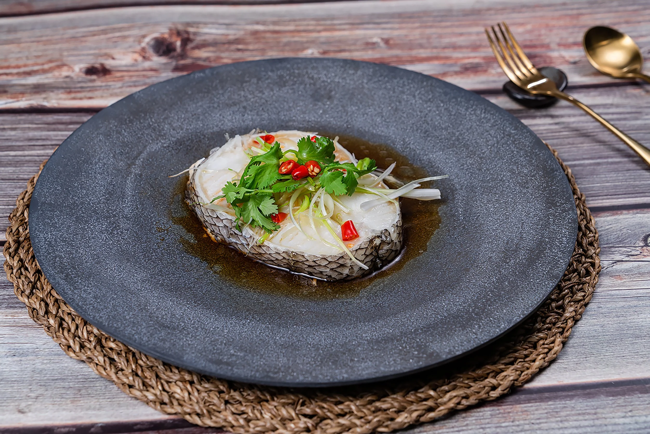 A cooked fish fillet garnished with cilantro and sliced red chili peppers, served on a dark plate with sauce, sitting on a woven placemat on a wooden table.