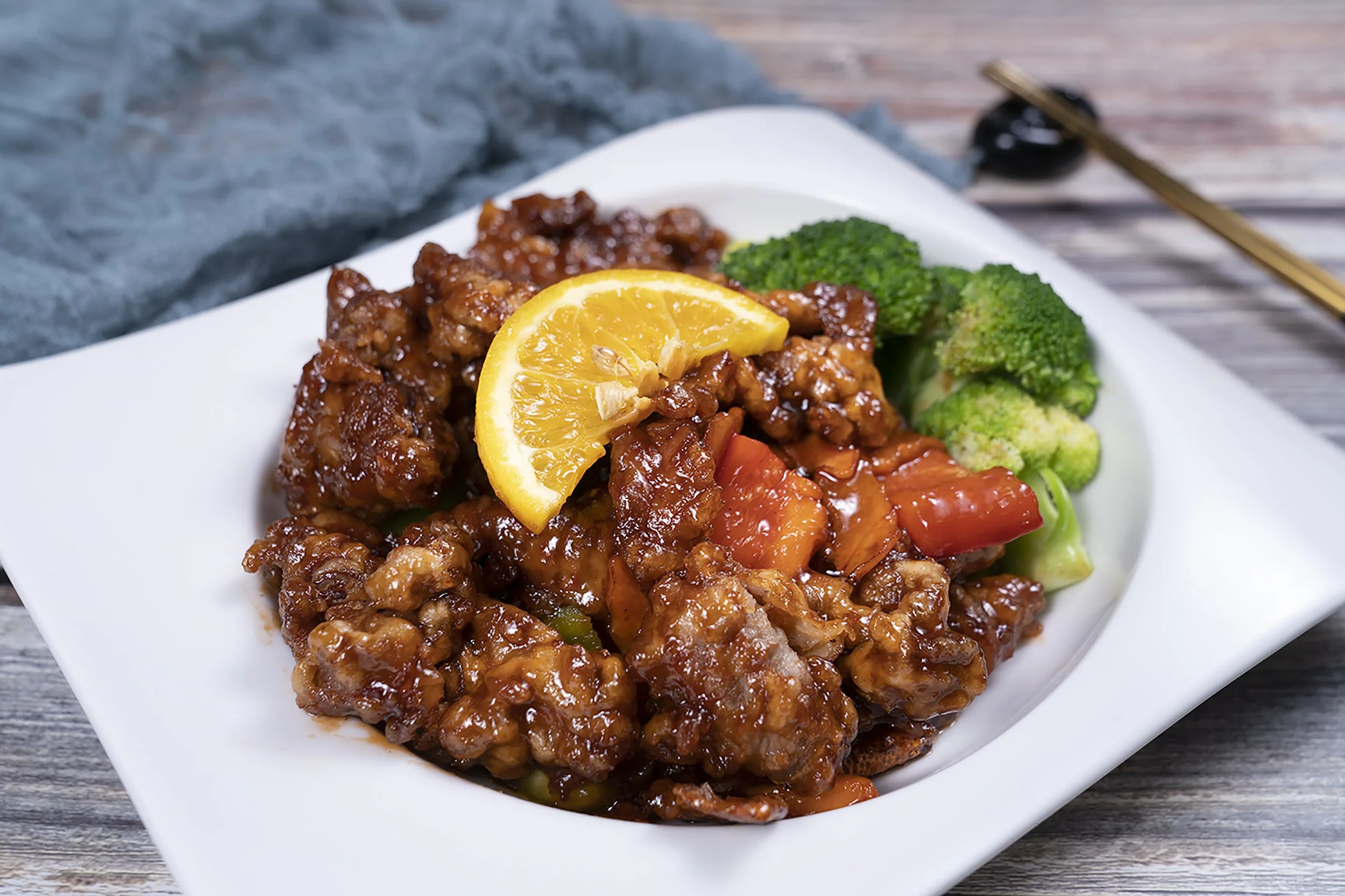 Close-up of a white plate containing sweet and sour chicken topped with a lemon wedge, served with broccoli and carrots on a wooden surface.