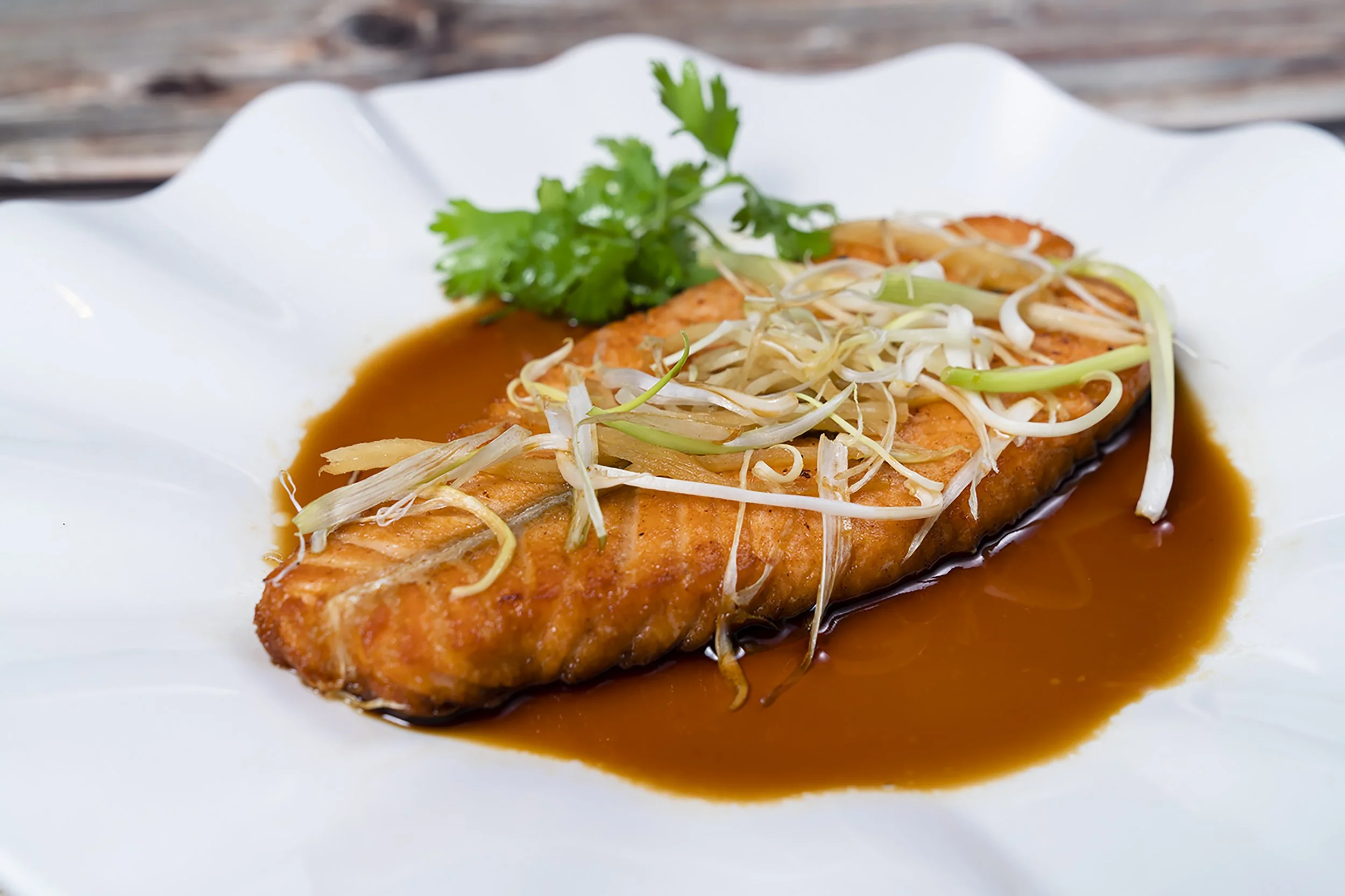 A cooked salmon fillet topped with shredded vegetables in sauce, garnished with cilantro, served on a white plate.
