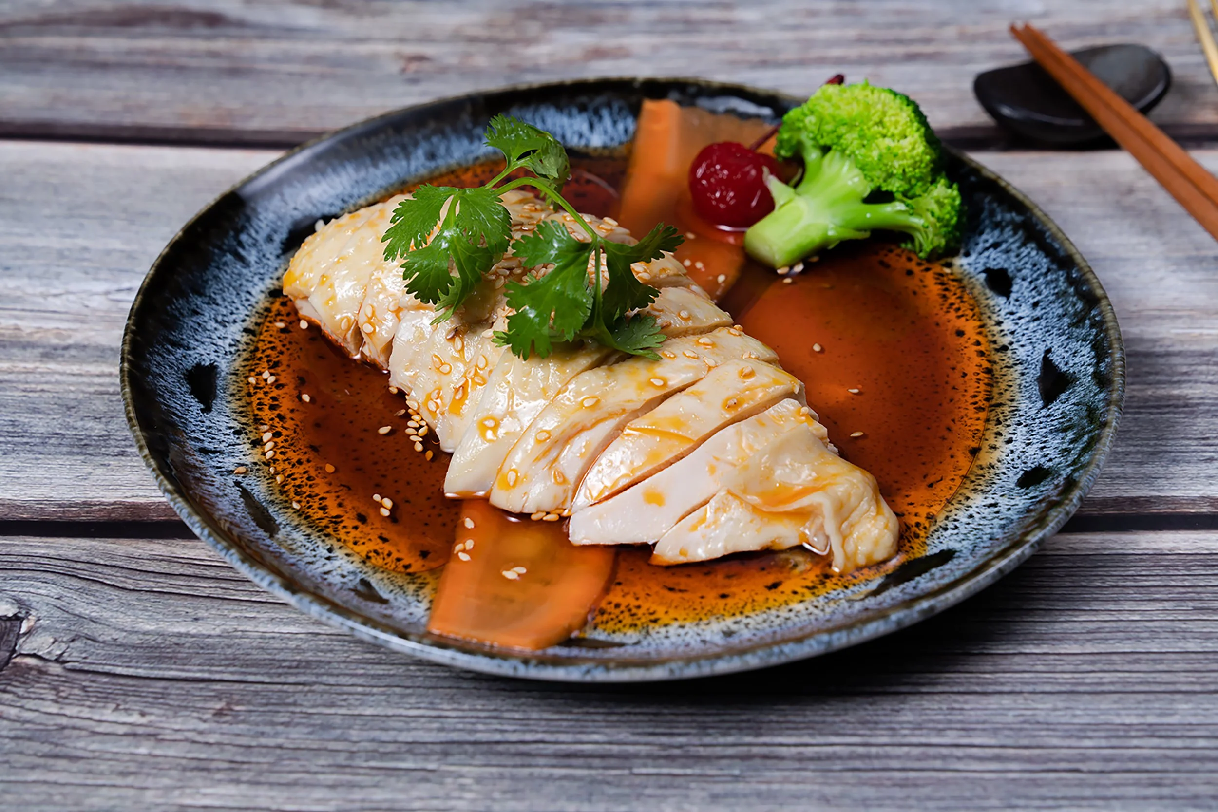 Sliced chicken breast on a black plate with sauce, topped with cilantro, alongside broccoli, a cherry, and a slice of vegetable, with a wooden surface background.