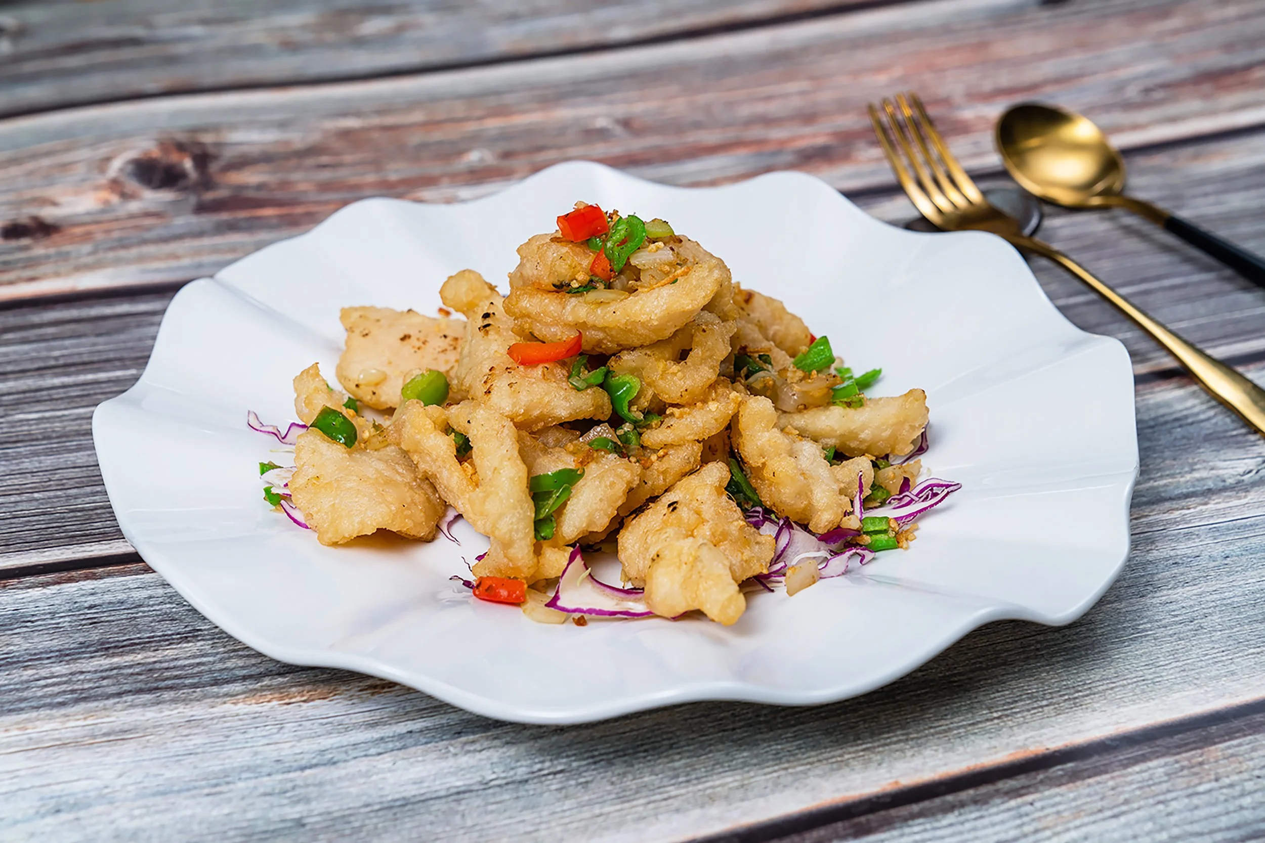 Fried fish pieces garnished with chopped green and red chilies on a white scalloped plate, placed on a rustic wooden table with a gold fork and spoon nearby.