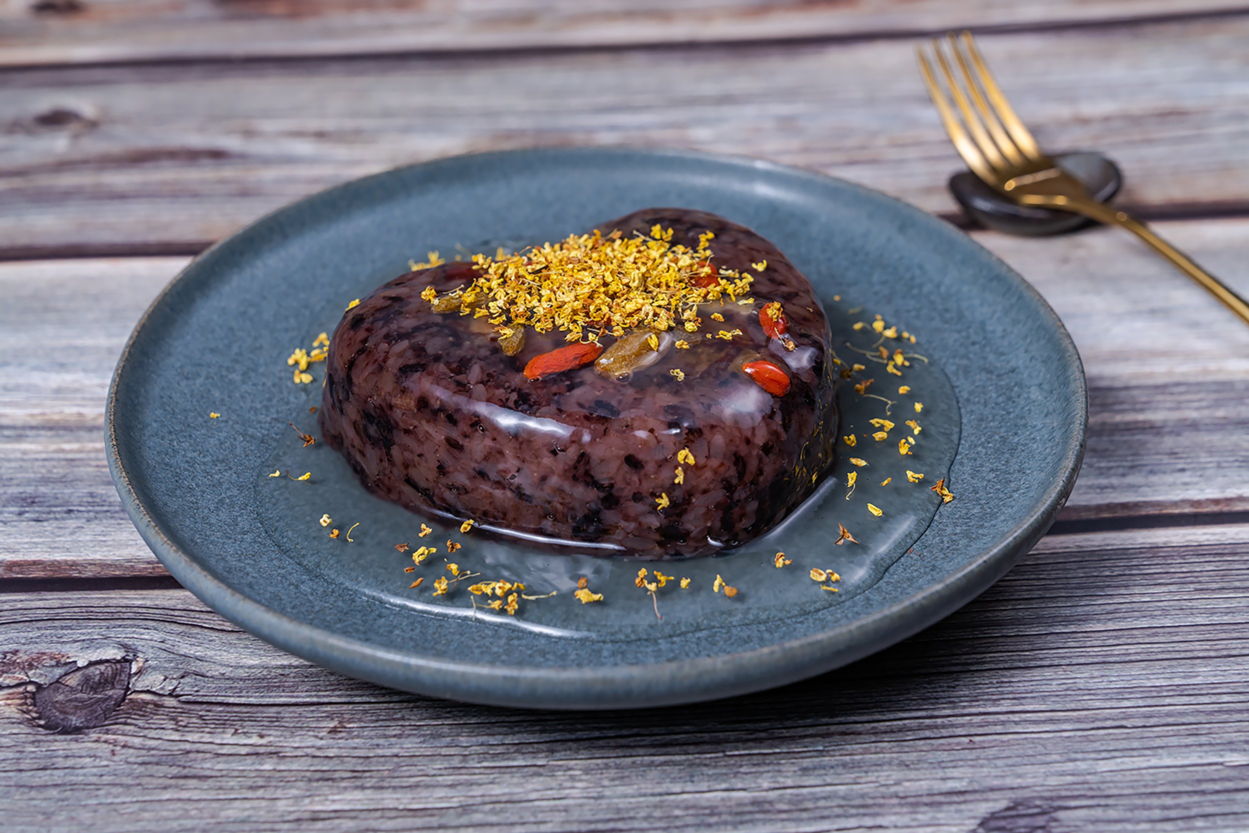 Chocolate dessert on a blue plate topped with yellow flower petals, placed on a wooden surface with a gold fork and black spoon nearby.