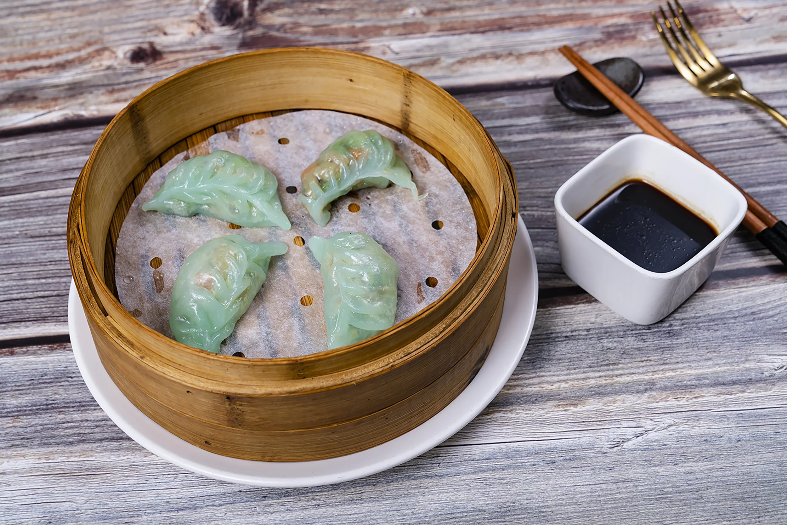 Steamed green dumplings in a bamboo steamer with soy sauce in a white square dish, chopsticks and a gold fork on a rustic wooden table.