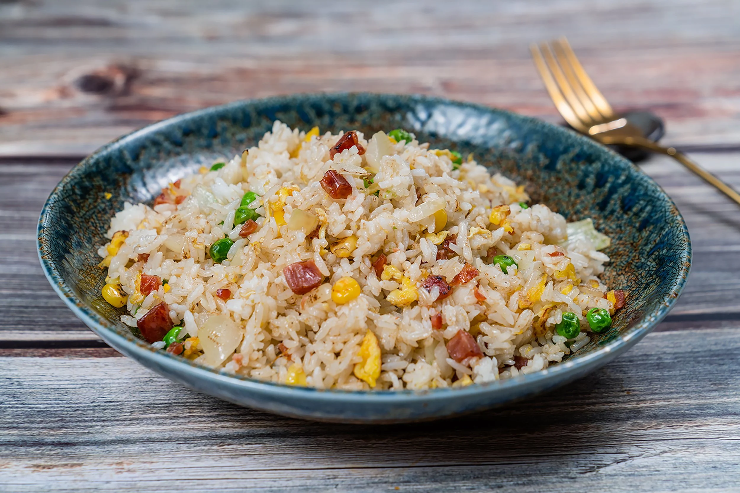 A bowl of fried rice with peas, diced ham, corn, and onions on a wooden table with a fork in the background.