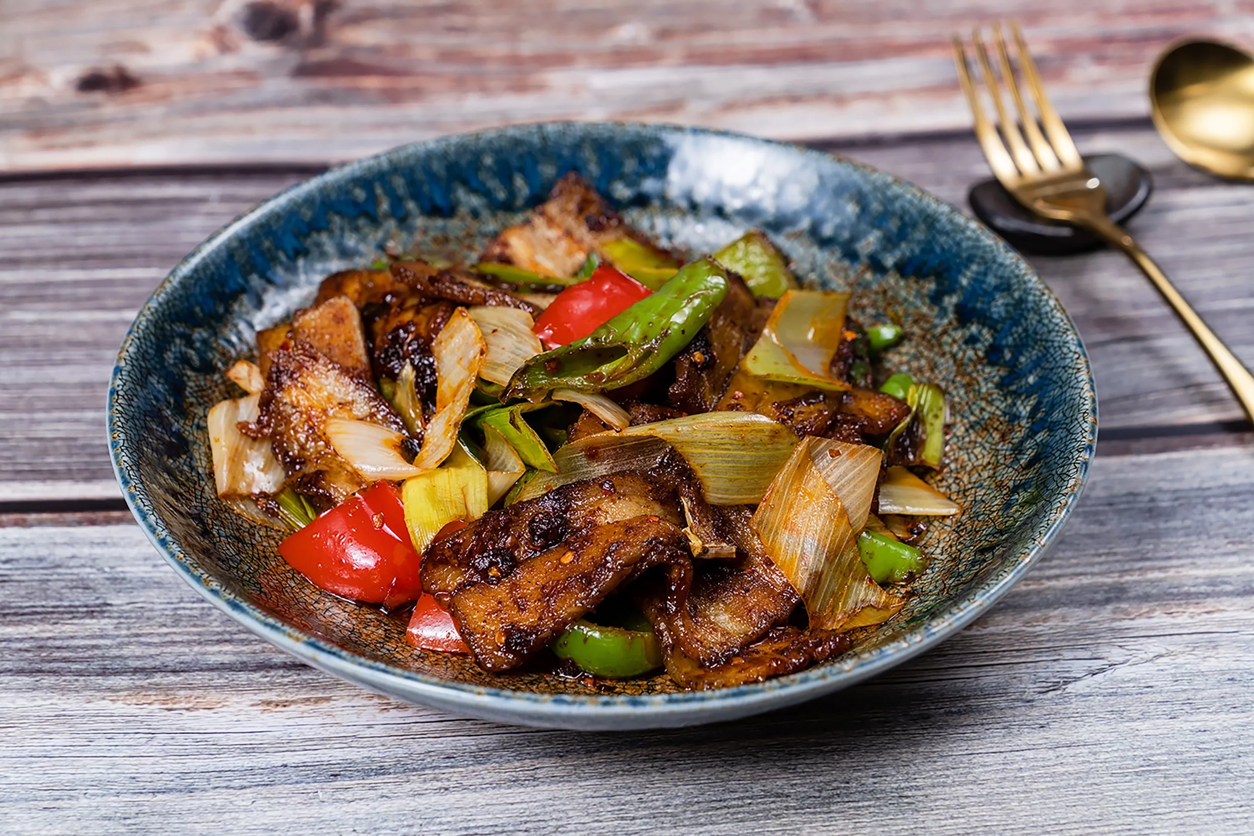 A bowl of stir-fried vegetables and meat with bell peppers, onions, and green beans on a wooden table, with a gold fork and spoon to the right.