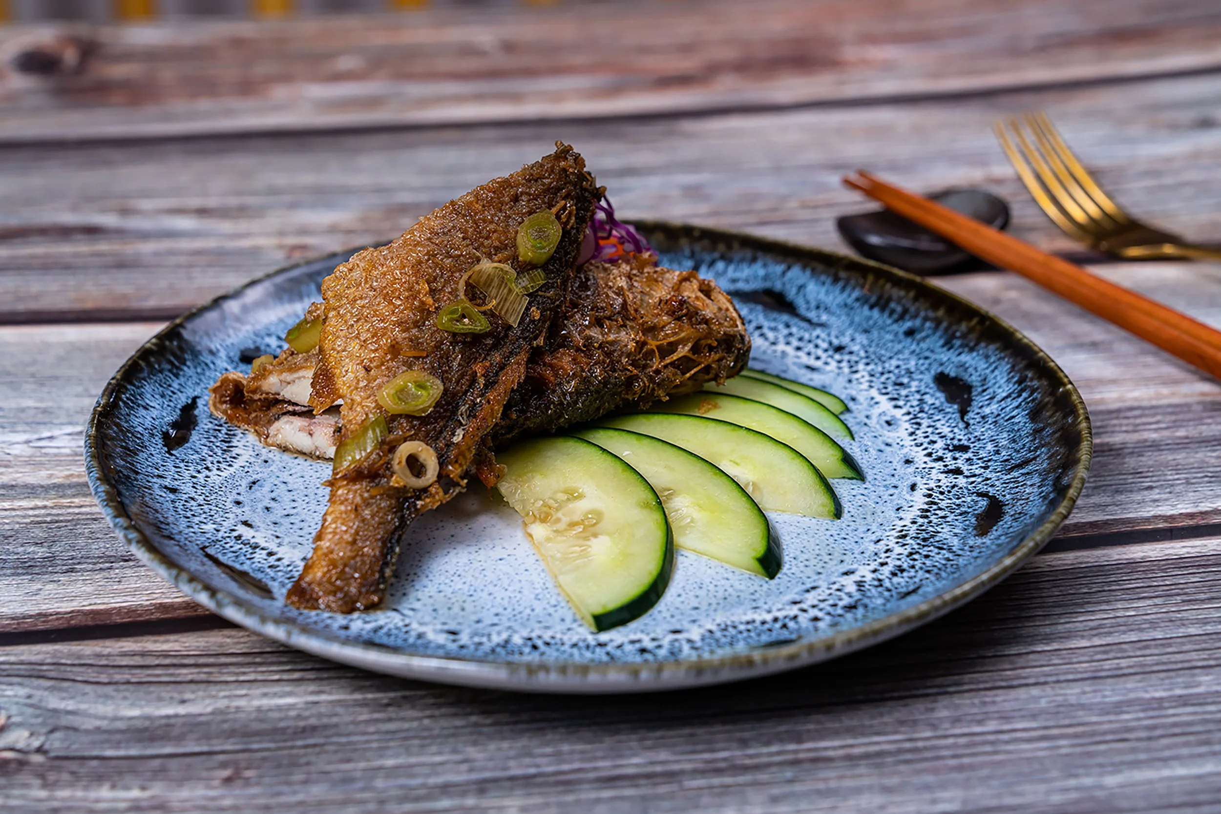 A plate with a cooked fish garnished with sliced green onions and served with sliced cucumber on a rustic wooden table.