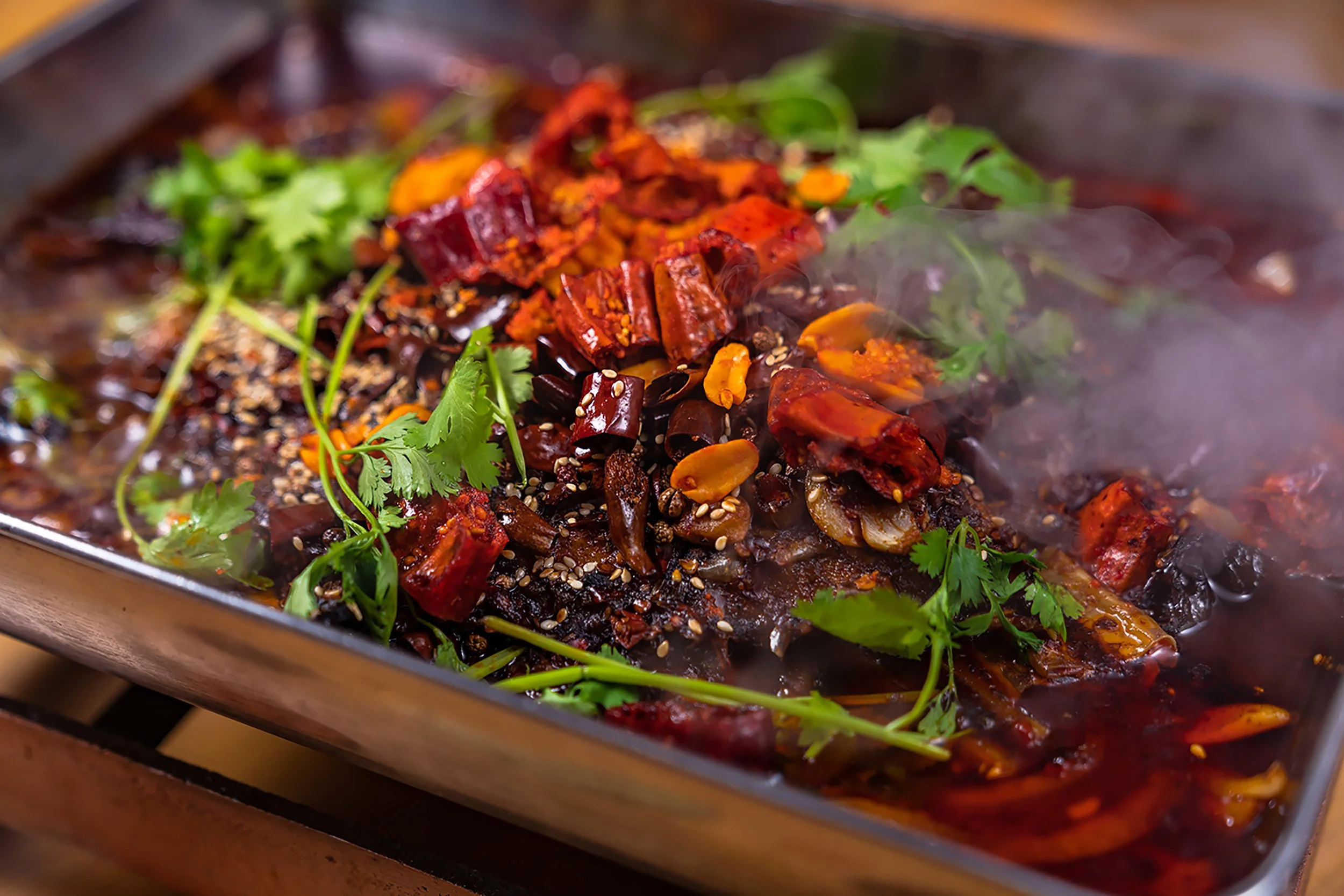 A steaming hot dish of spicy roasted meat garnished with fresh cilantro and sesame seeds, with a rich sauce underneath.
