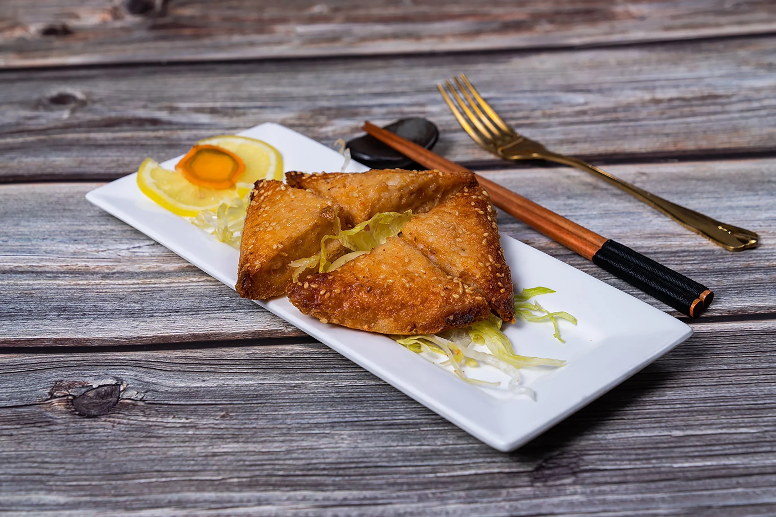 Fried fish fillets with lemon slices on a white rectangular plate, set on a rustic wooden surface, with chopsticks and a spoon nearby.