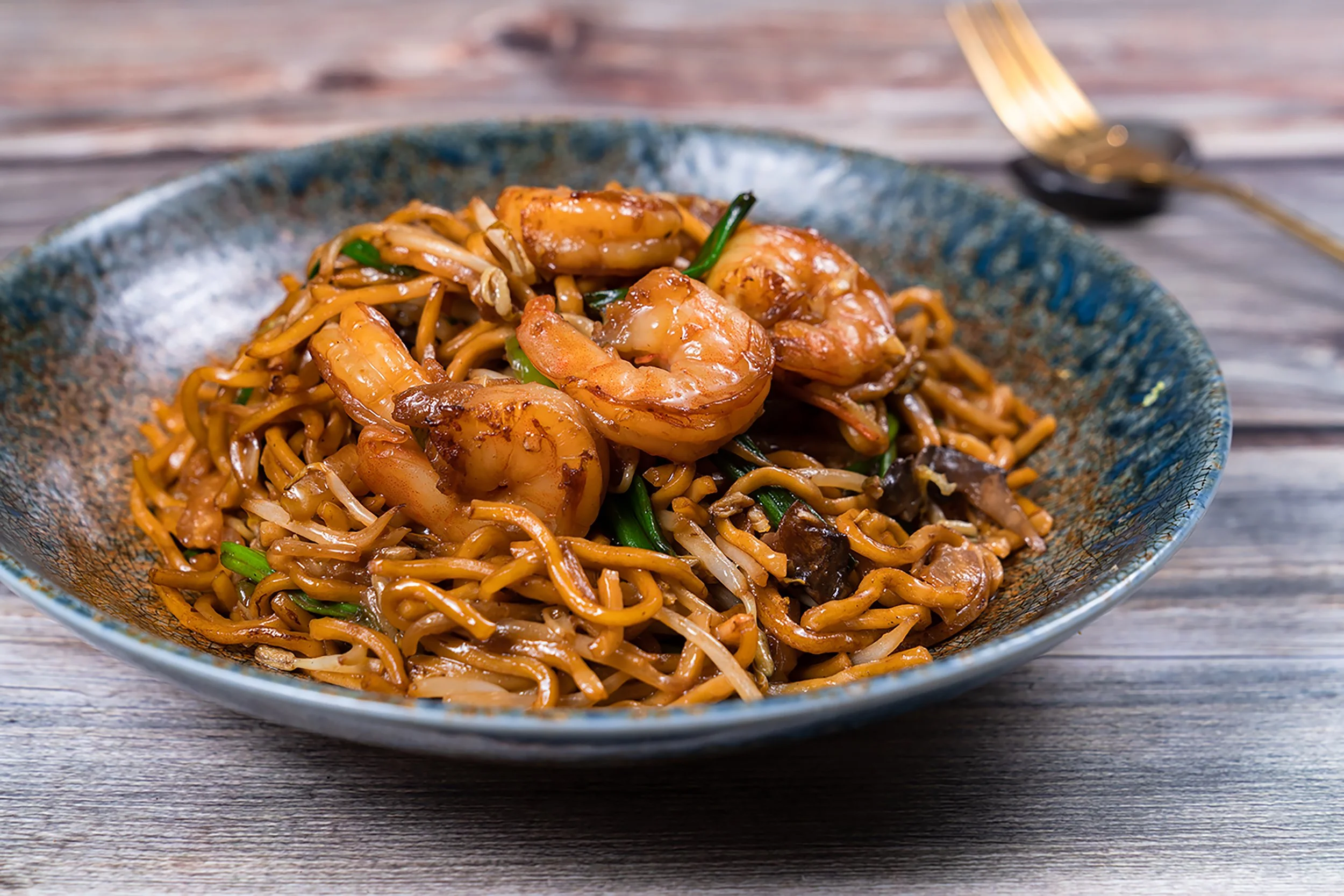 A bowl of stir-fried noodles with shrimp, green onions, and vegetables on a wooden table.