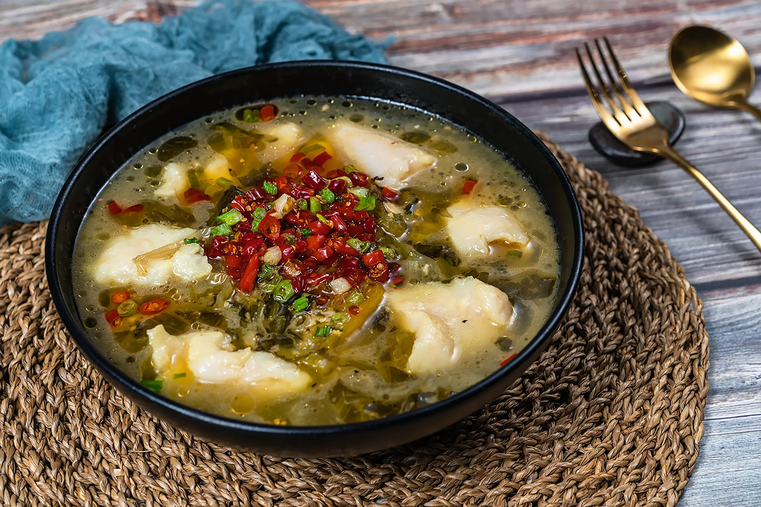 A bowl of fish soup topped with chopped red and green chilies on a woven placemat on a rustic wooden table, with a gold fork and spoon beside.