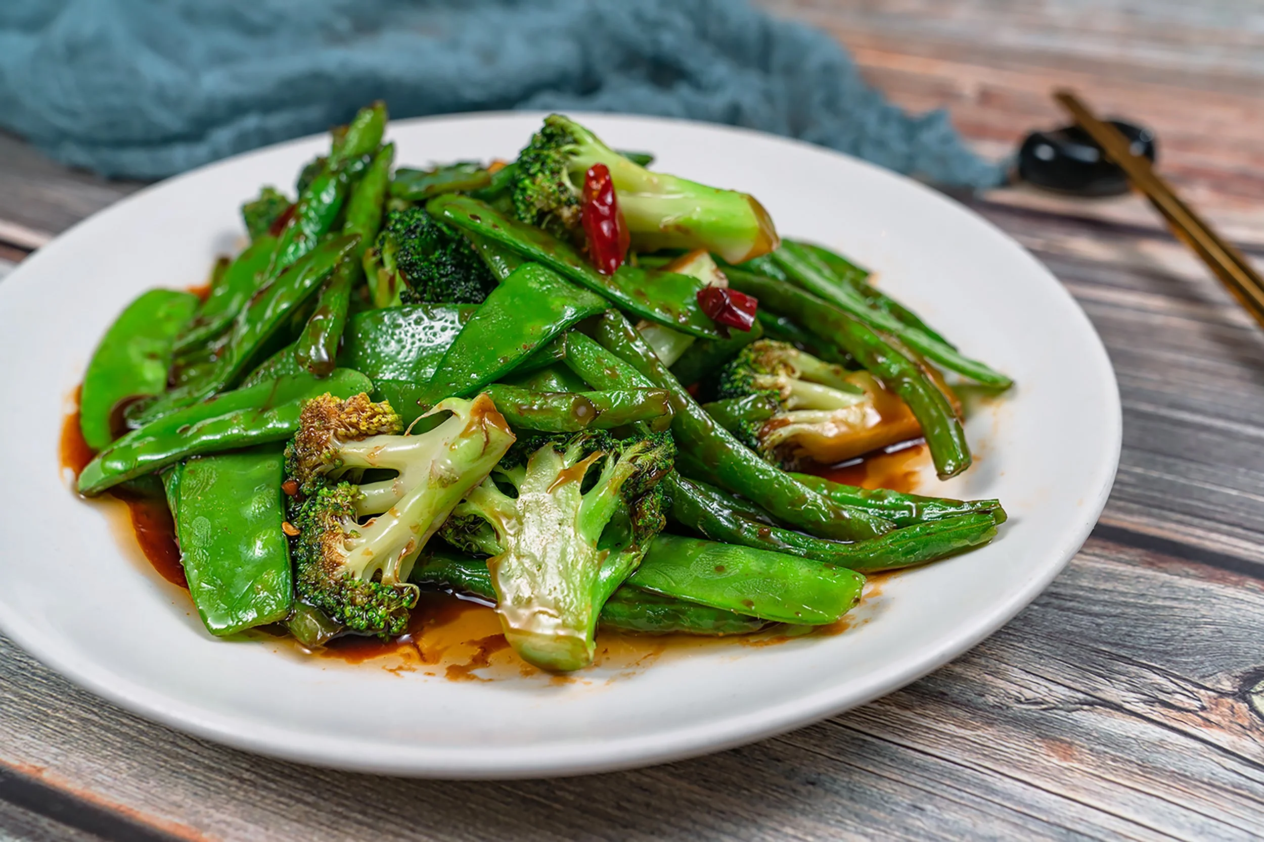 A white plate filled with stir-fried green vegetables including broccoli, snap peas, and possibly bok choy, with a brown sauce on top, placed on a wooden table.