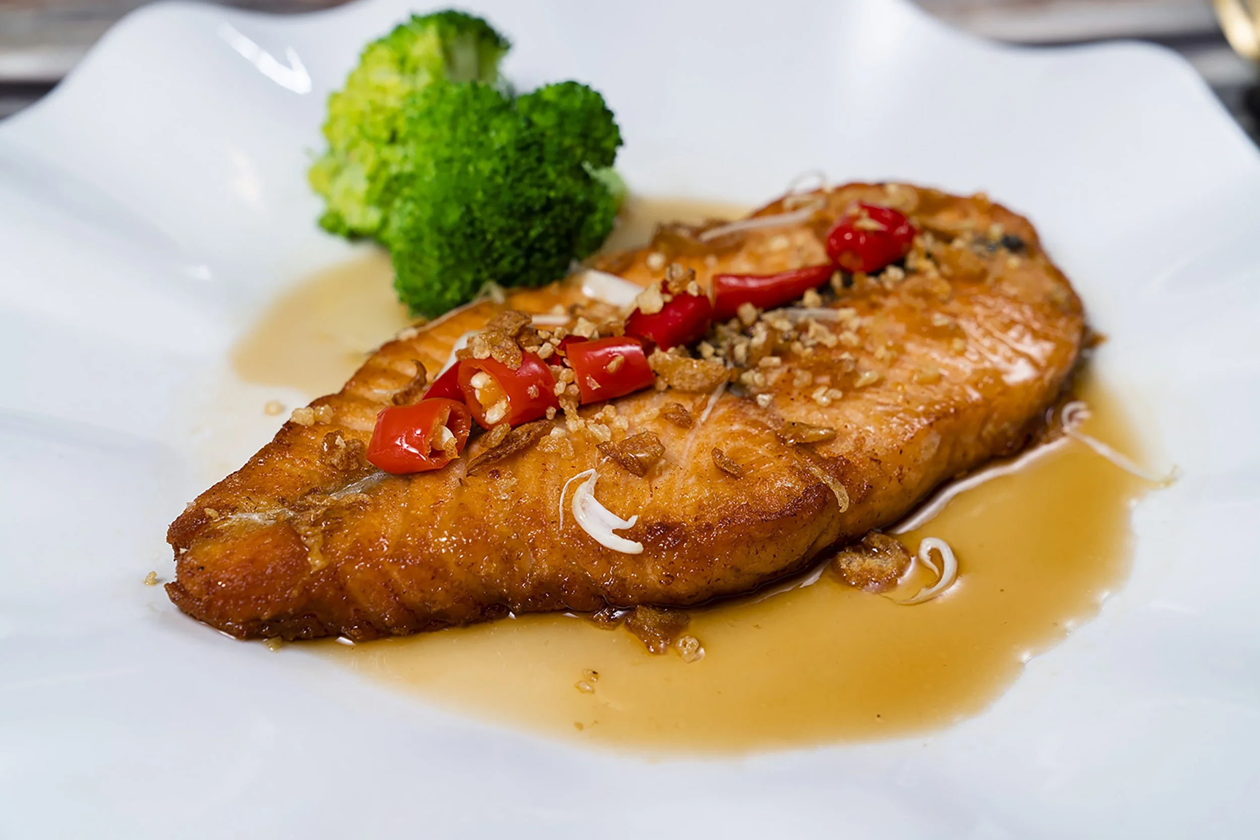 A plated dish featuring a seasoned, cooked salmon fillet topped with chopped red peppers and chopped nuts, served with a side of steamed broccoli on a white plate.