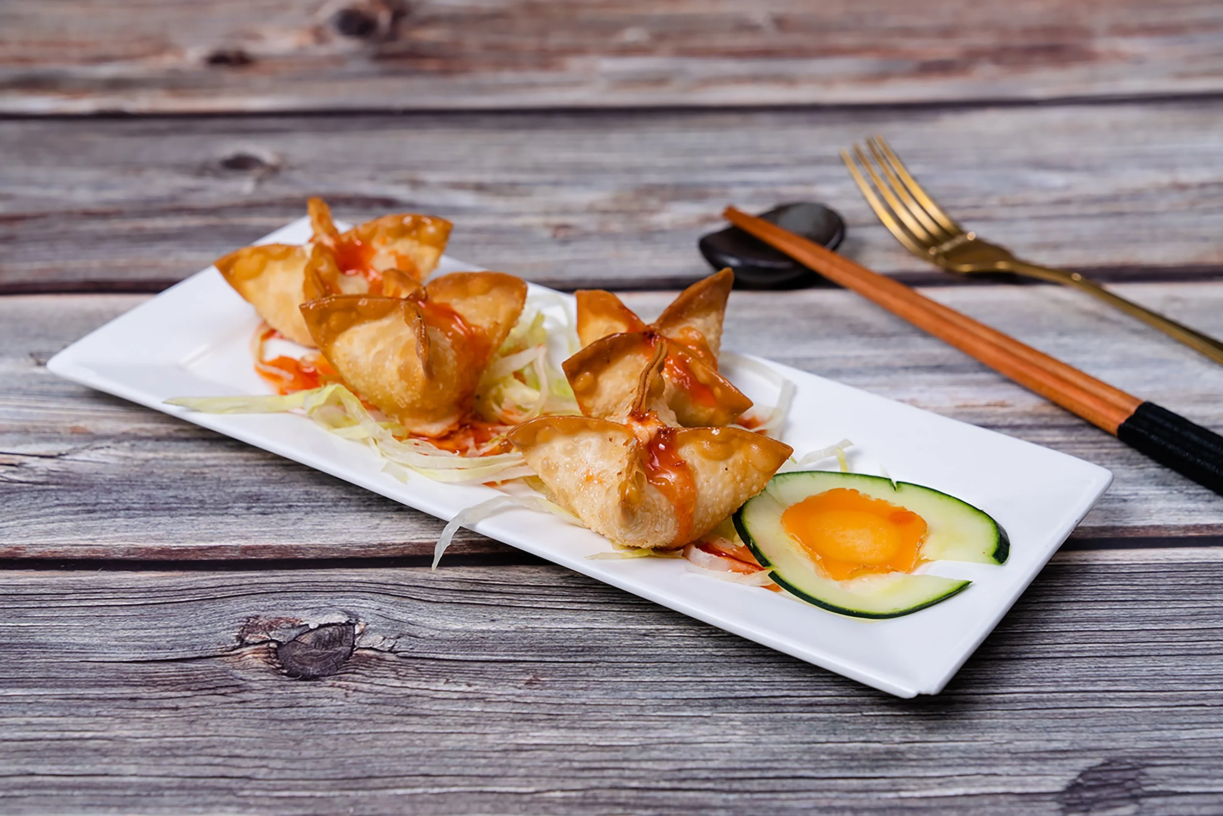 Fried wontons drizzled with sauce served on a white rectangular plate with shredded lettuce and cucumber slices, on a rustic wooden table.