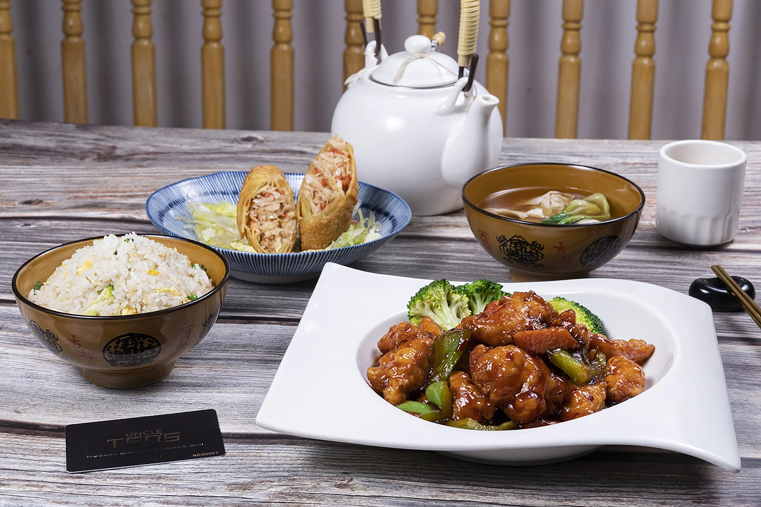 A variety of Asian dishes on a wooden table, including fried spring rolls filled with vegetables and meat, fried rice, a bowl of soup with vegetables, and an orange chicken dish with broccoli and carrots. There is also a teapot and a white mug in the background.