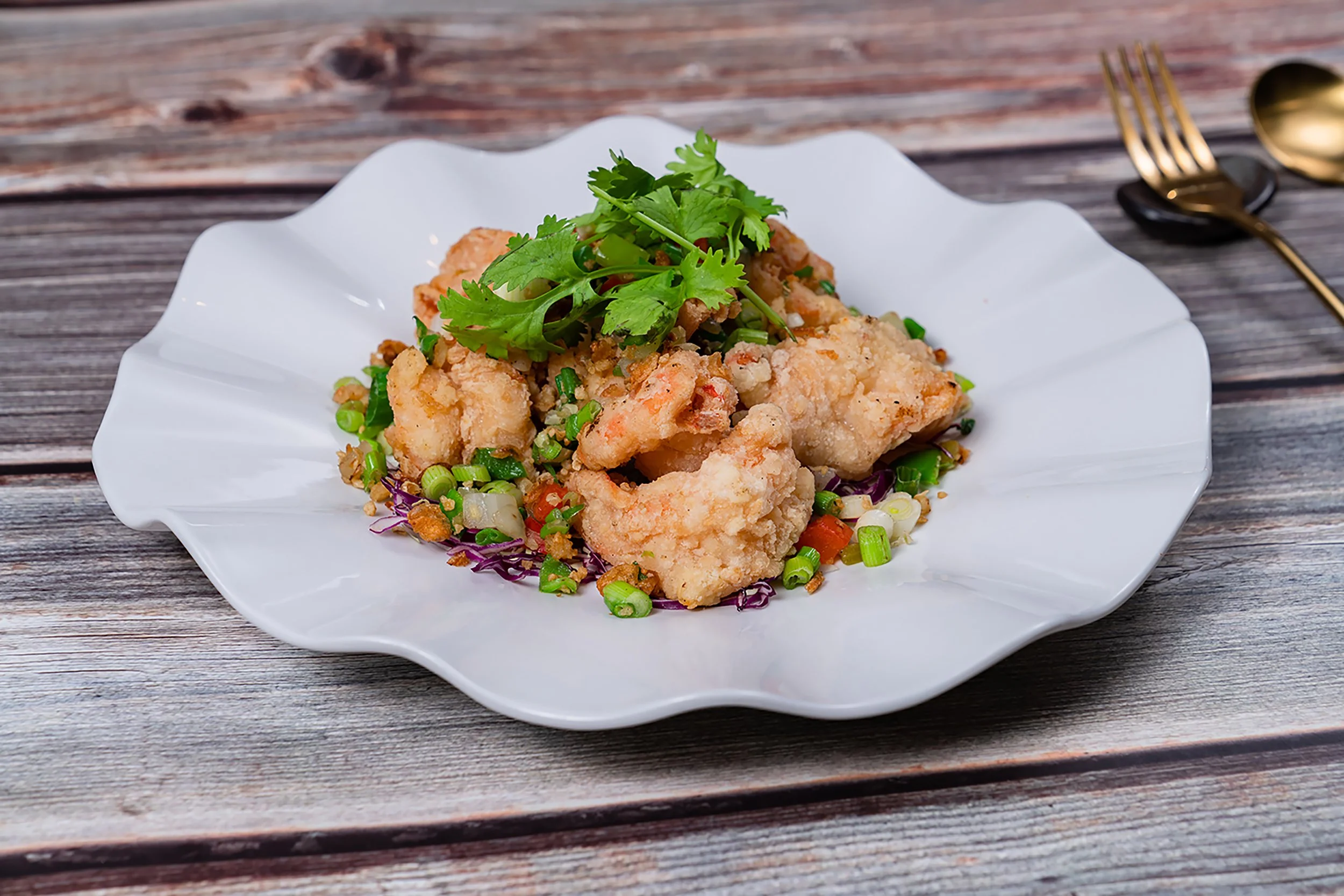 Fried fish fillet topped with fresh cilantro on a white decorative plate, accompanied by chopped green onions and vegetables, on a rustic wooden table with a gold spoon and fork to the side.