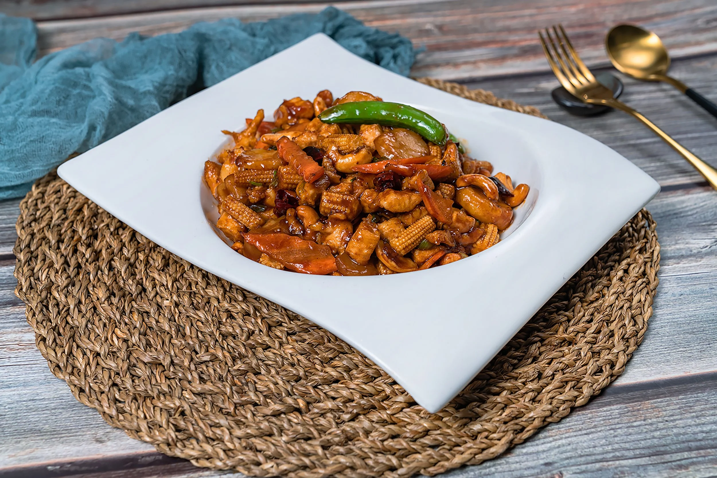 A plate of stir-fried vegetables and chicken with a green chili on top, served on a square white dish, resting on a woven placemat on a wooden table.