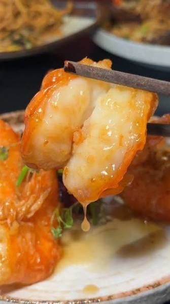 A close-up of a piece of cooked seafood, likely shrimp, held by chopsticks with some sauce dripping from it. In the background, there are other dishes or bowls.