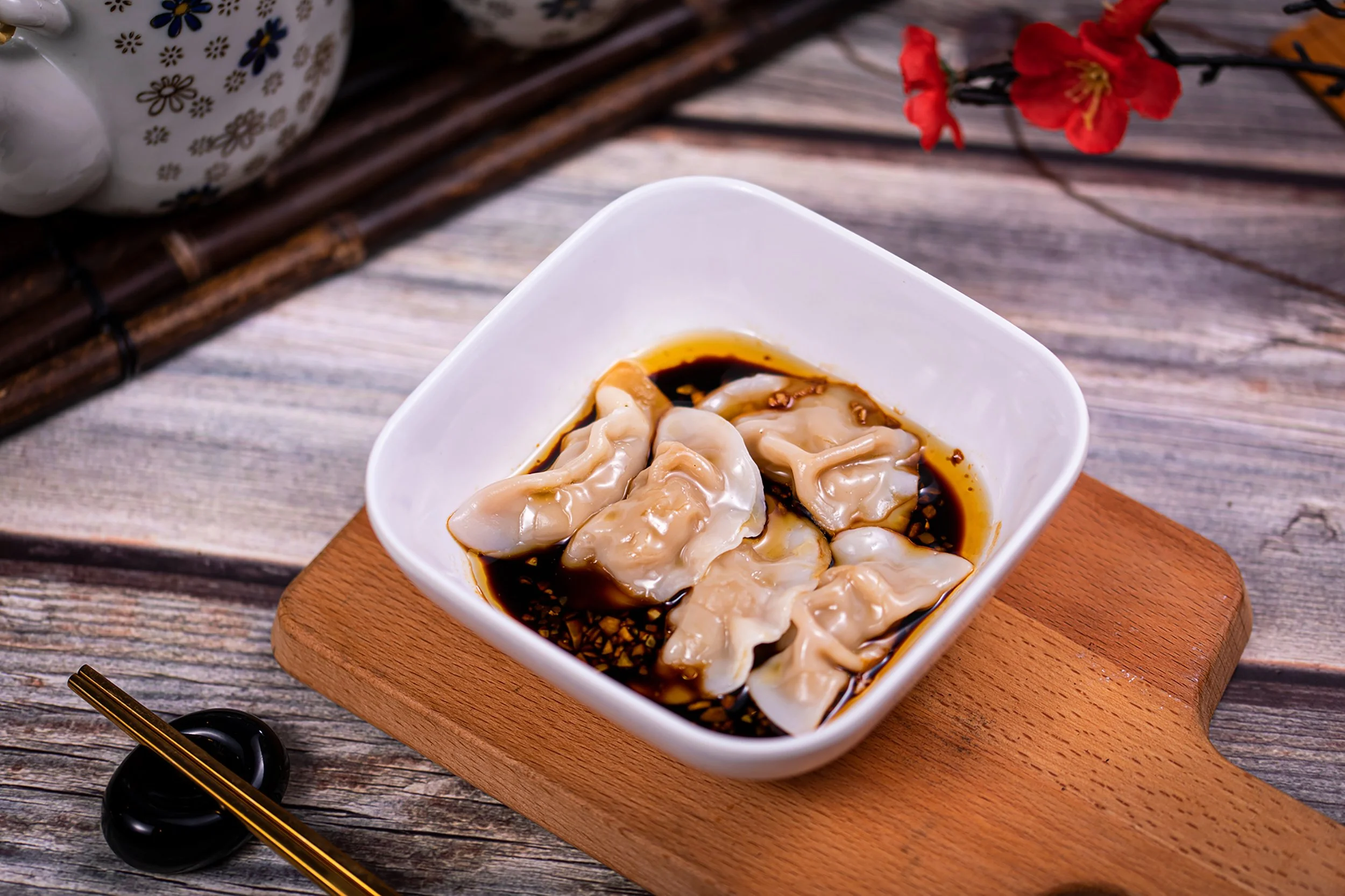 Steamed dumplings in a soy sauce-based dipping sauce served in a white square bowl on a wooden tray, with chopsticks and a black spoon nearby, set on a rustic wooden table with a teapot and red flower in the background.
