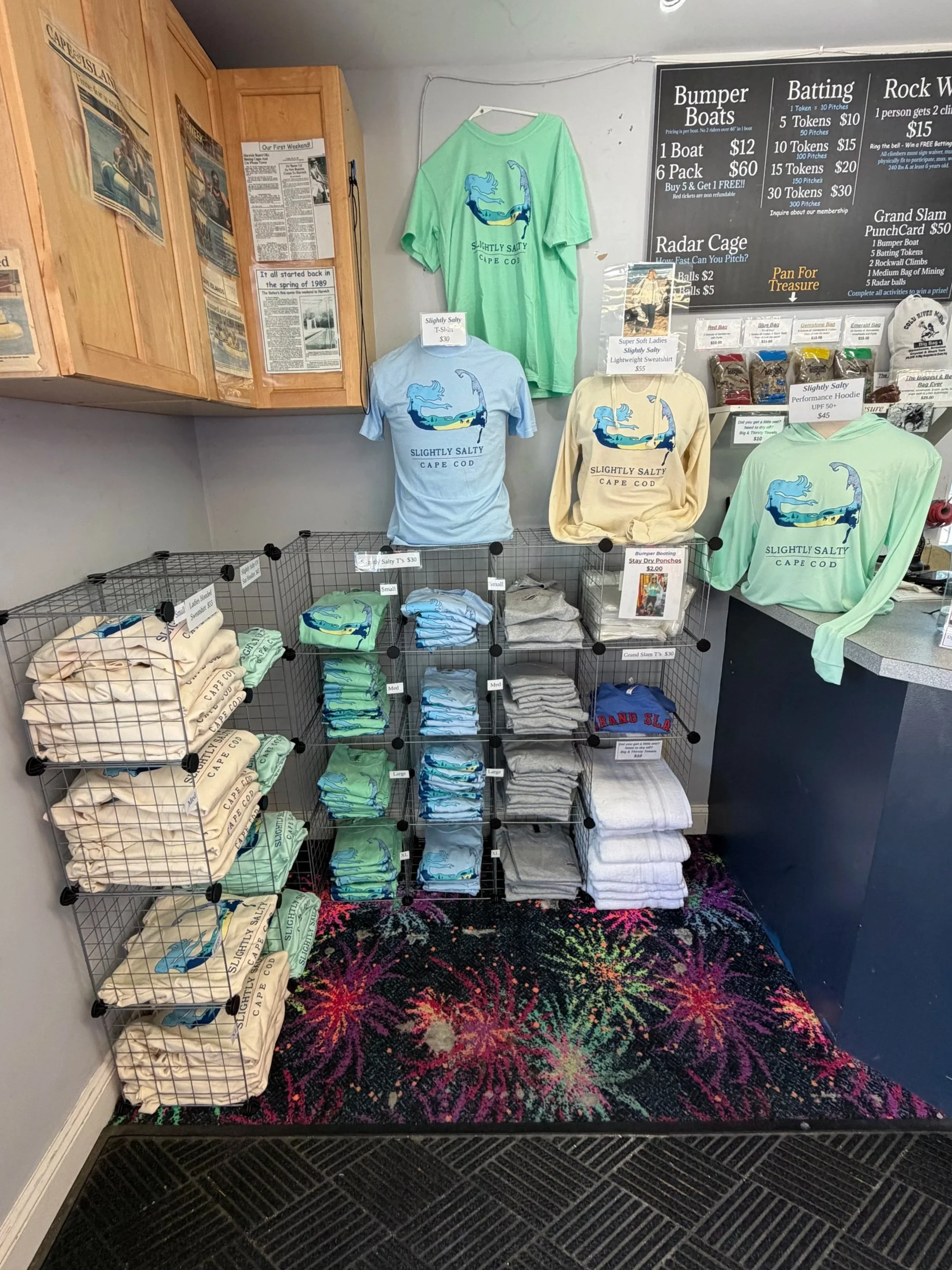 Display of colorful t-shirts, hoodies, and folded clothing items on metal racks and shelves inside a store. A menu board with prices and a black floor mat with a floral pattern is visible at the bottom.