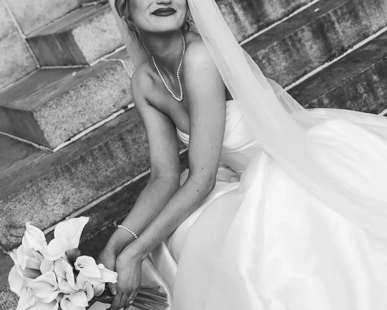 Black and white photo of a bride sitting on stone steps, wearing a strapless wedding gown, veil, and pearl jewelry, holding a bouquet of flowers.