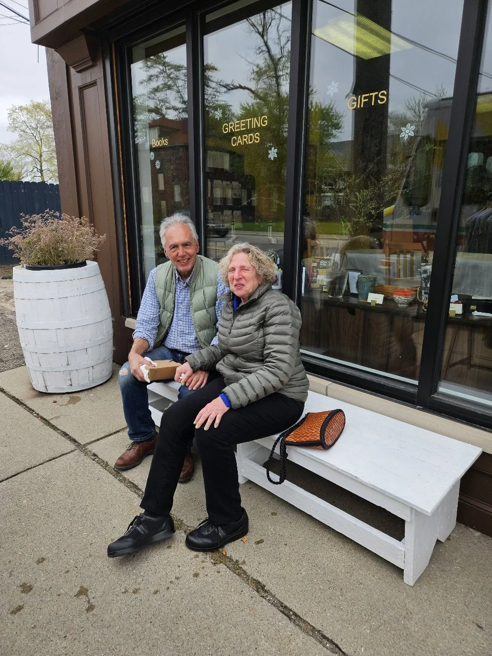 Two smiling seniors sitting on a white wooden bench outside a storefront with large glass windows, one man and one woman, holding a small gift box, with potted plant nearby and signs on glass advertising books, greeting cards, and gifts.
