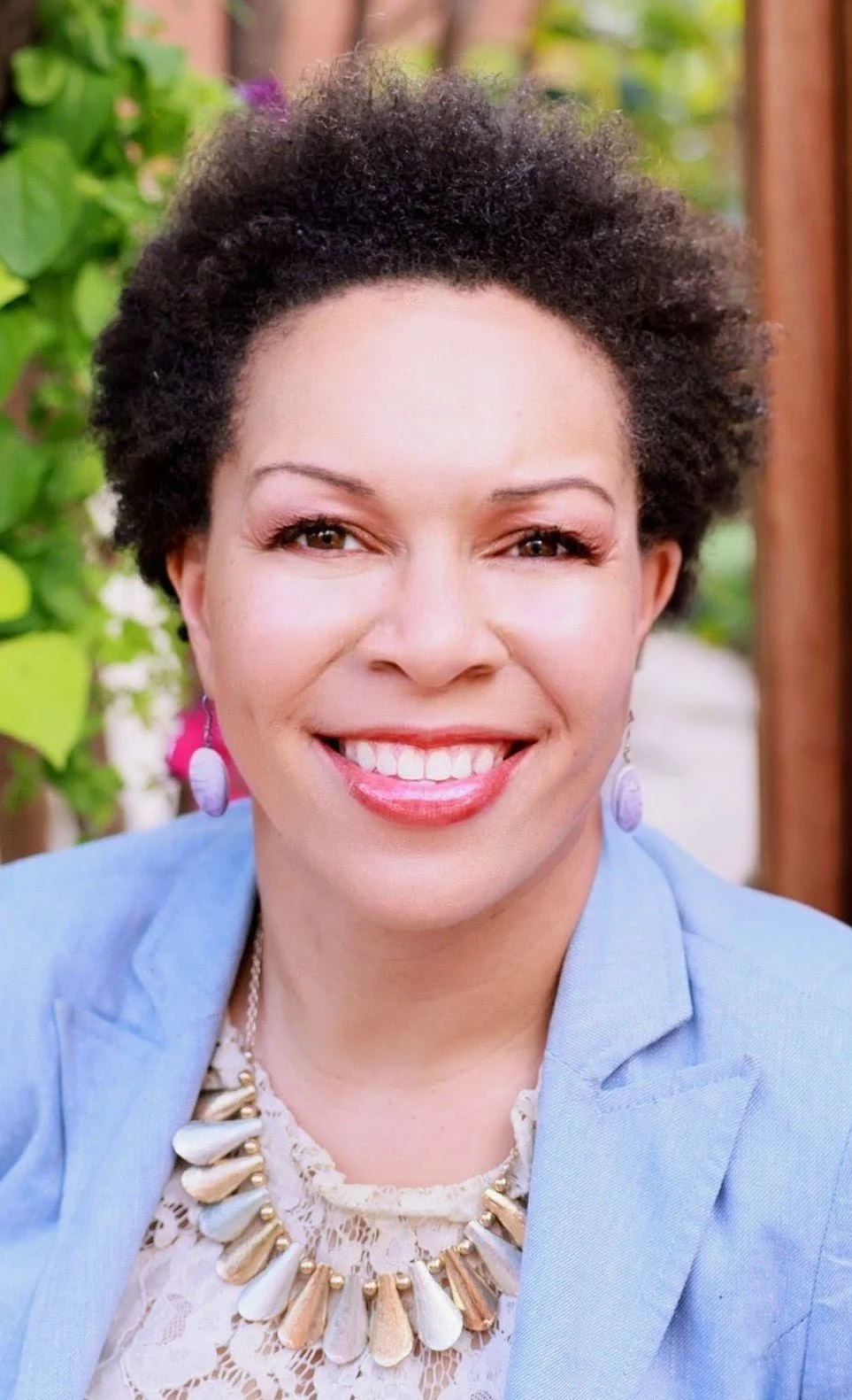 A smiling woman with short, curly dark hair, wearing a light blue blazer, a lace top, a statement necklace with large beige and white beads, and pink earrings, outdoors with green foliage in the background.