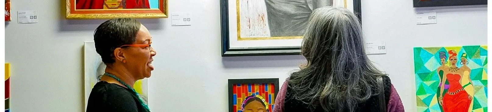 Two women are looking at colorful artwork displayed on a gallery wall. One woman has short hair, glasses, and is smiling, while the other woman has long gray hair and is facing the artworks.