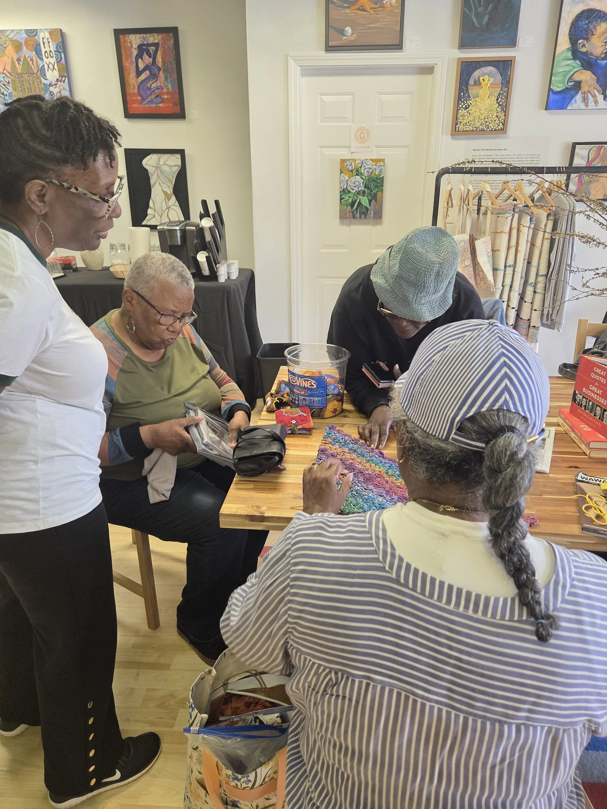 A group of women gathered inside an art gallery, engaging in a craft or conversation around a wooden table with various art supplies and colorful fabric. Several paintings hang on the walls.