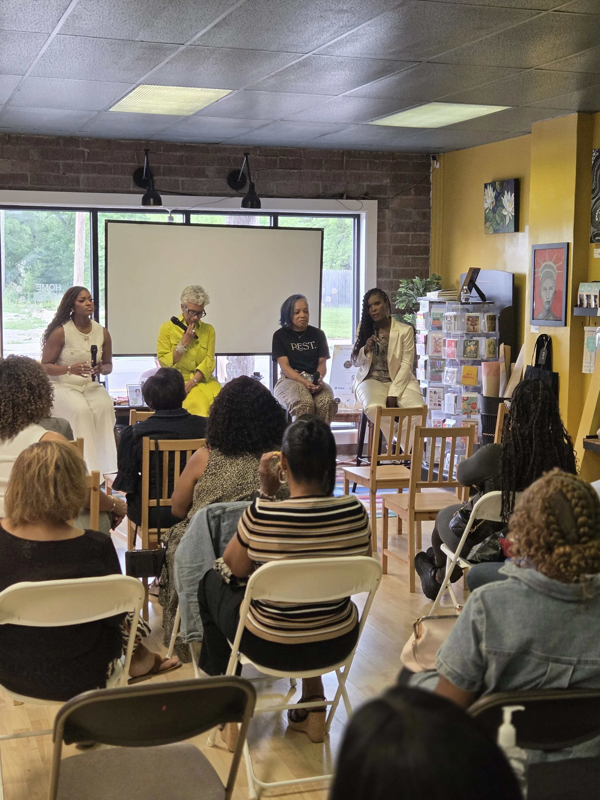 A panel of four women speaking at an indoor event, seated on a stage in front of an audience. The room has a brick wall, large window, and colorful artwork on the walls.