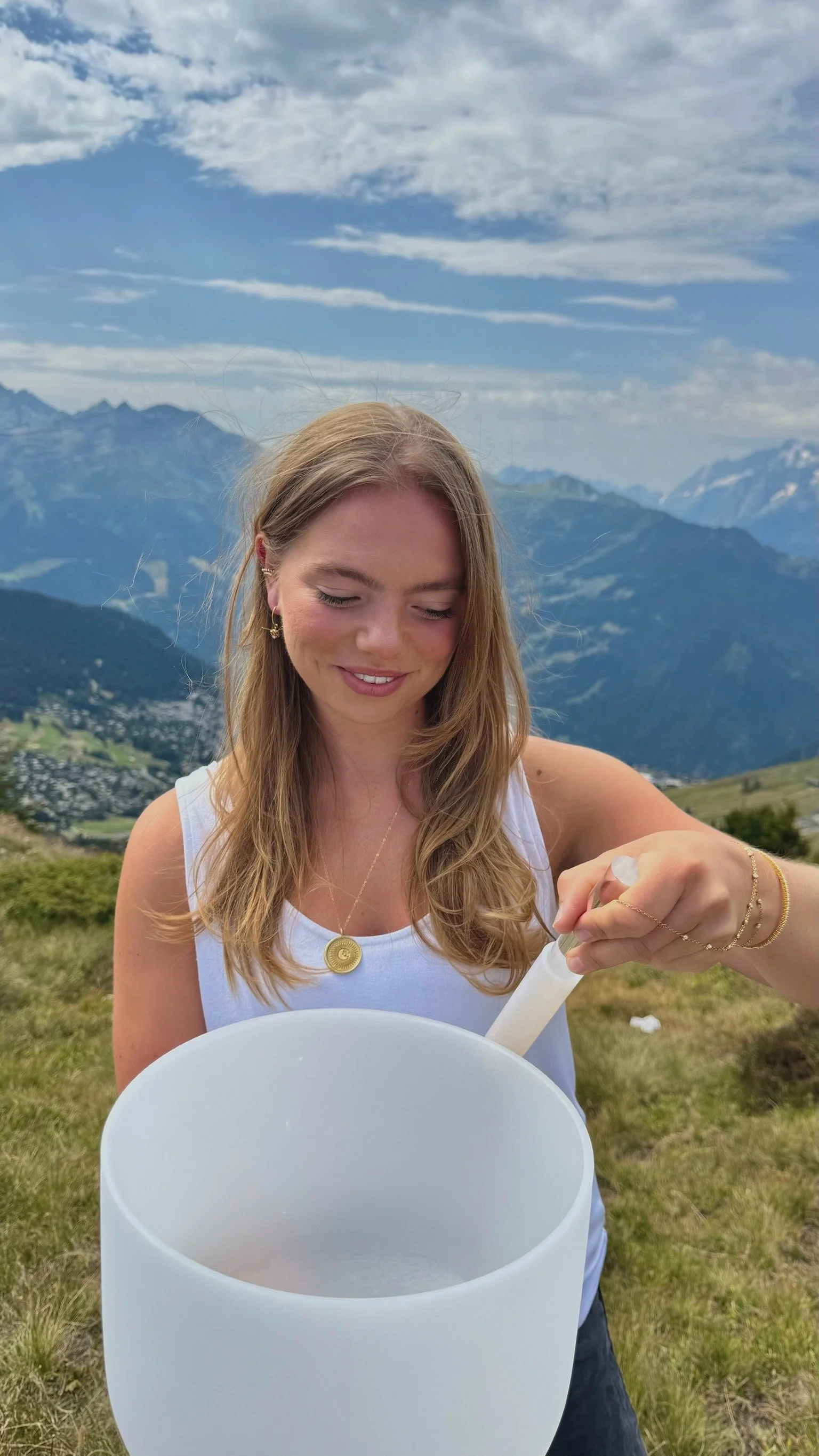A young woman with long blonde hair, smiling with her eyes closed, holds a crystal singing bowl and stands outdoors in a mountain landscape with blue sky, clouds, and distant mountains in the background.