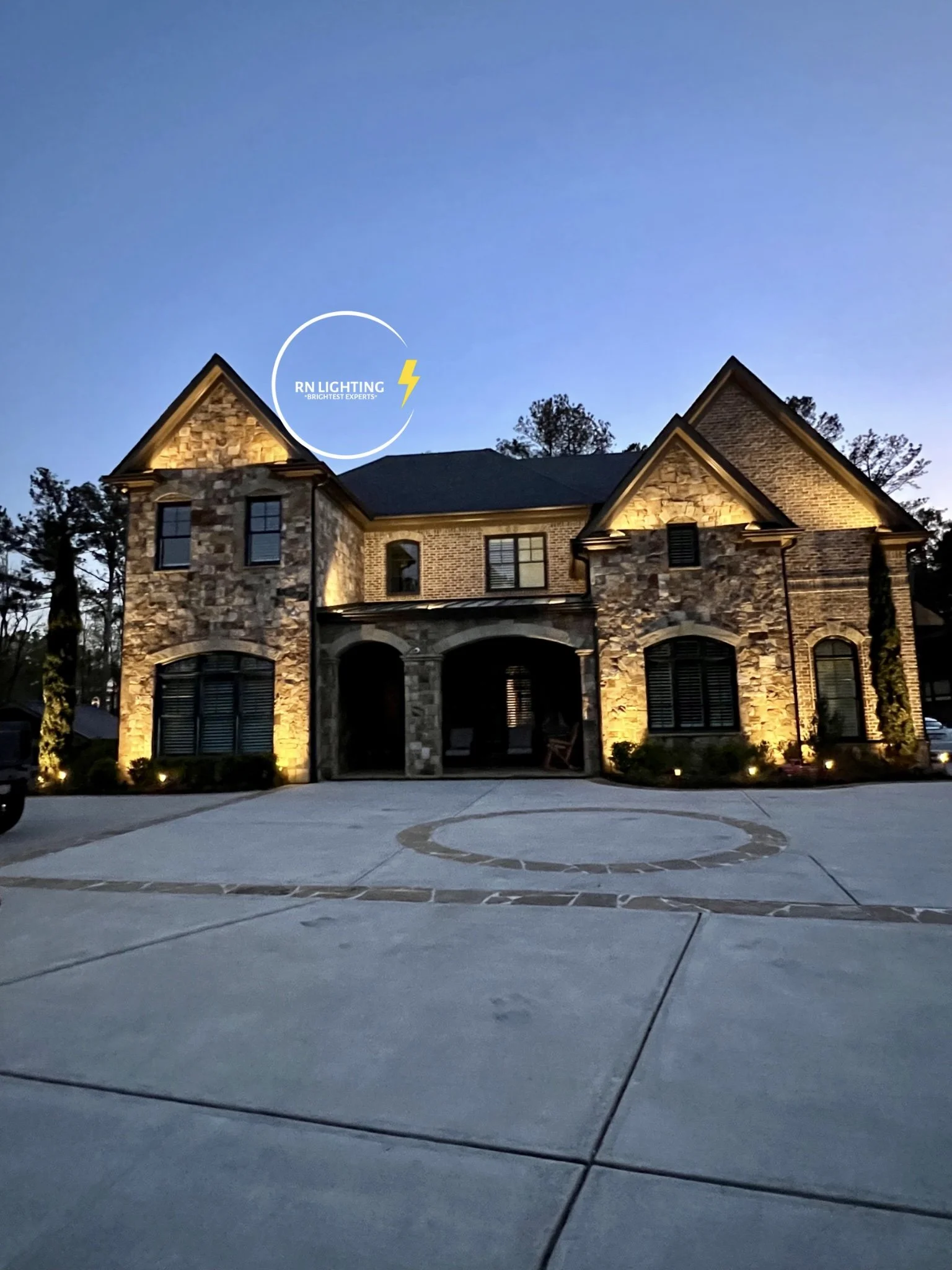 A large brick and stone house with exterior lighting illuminating its facade, views of trees and a driveway in front.