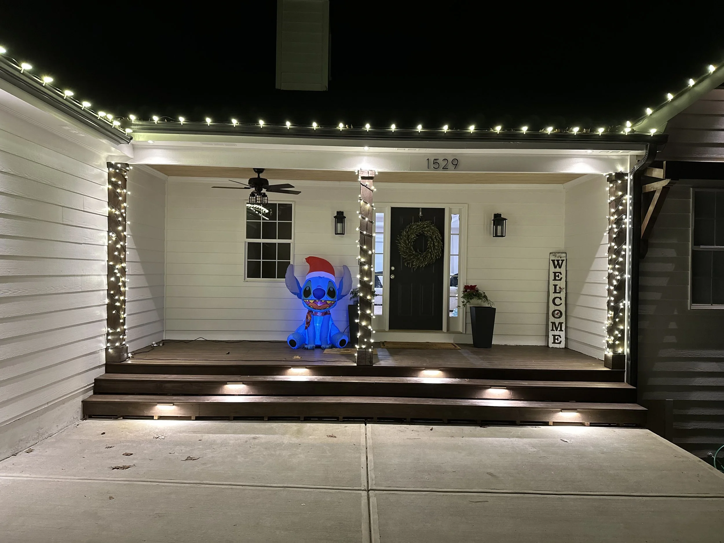 Night view of a house front porch decorated with string lights, a welcome sign, a plant, and an inflatable Stitch character wearing a Santa hat.