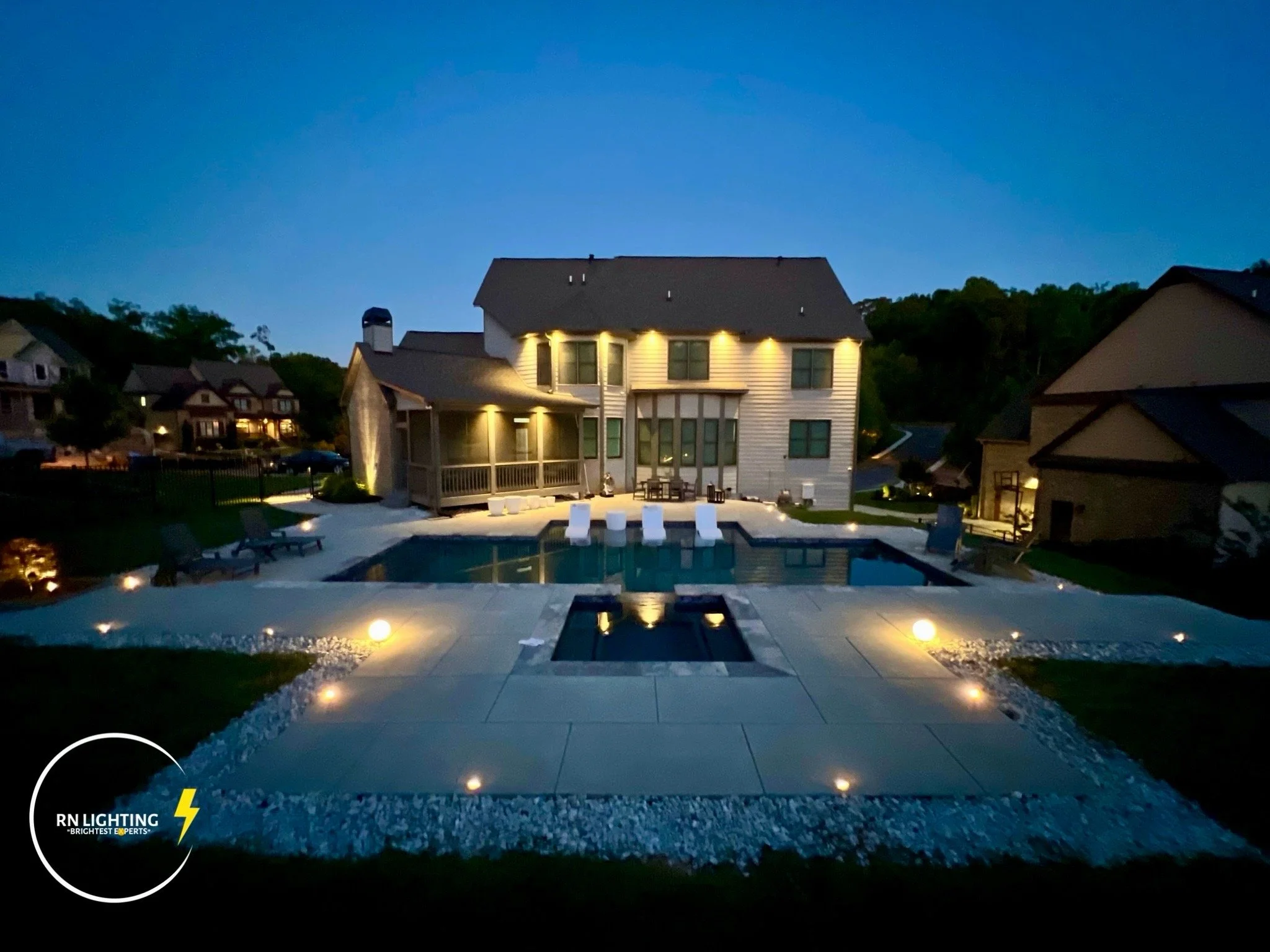 Nighttime view of a house with exterior lights, a swimming pool, and a backyard patio with seating. The house is surrounded by other houses and trees, under a dark sky.