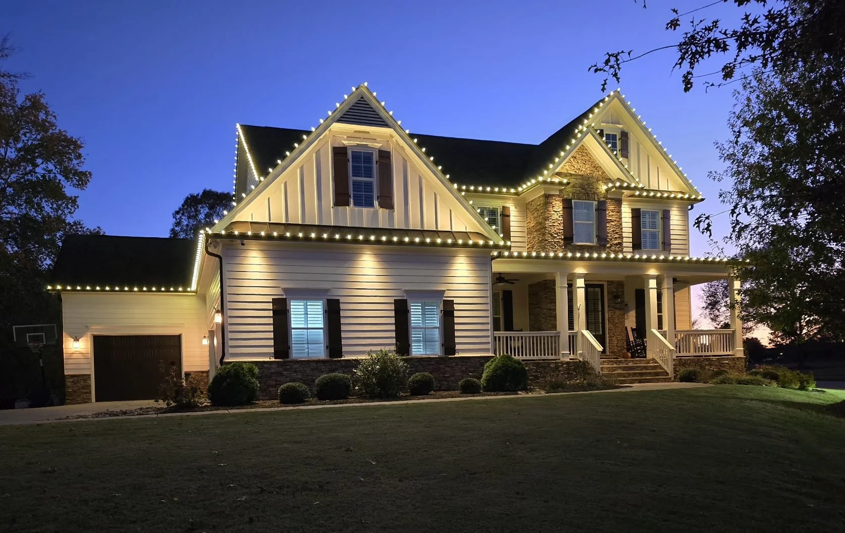 A large two-story house with decorative white lights outlining the roof and porch, featuring a front porch with white columns, multiple windows with shutters, a garage, and a well-kept lawn during sunset or twilight.