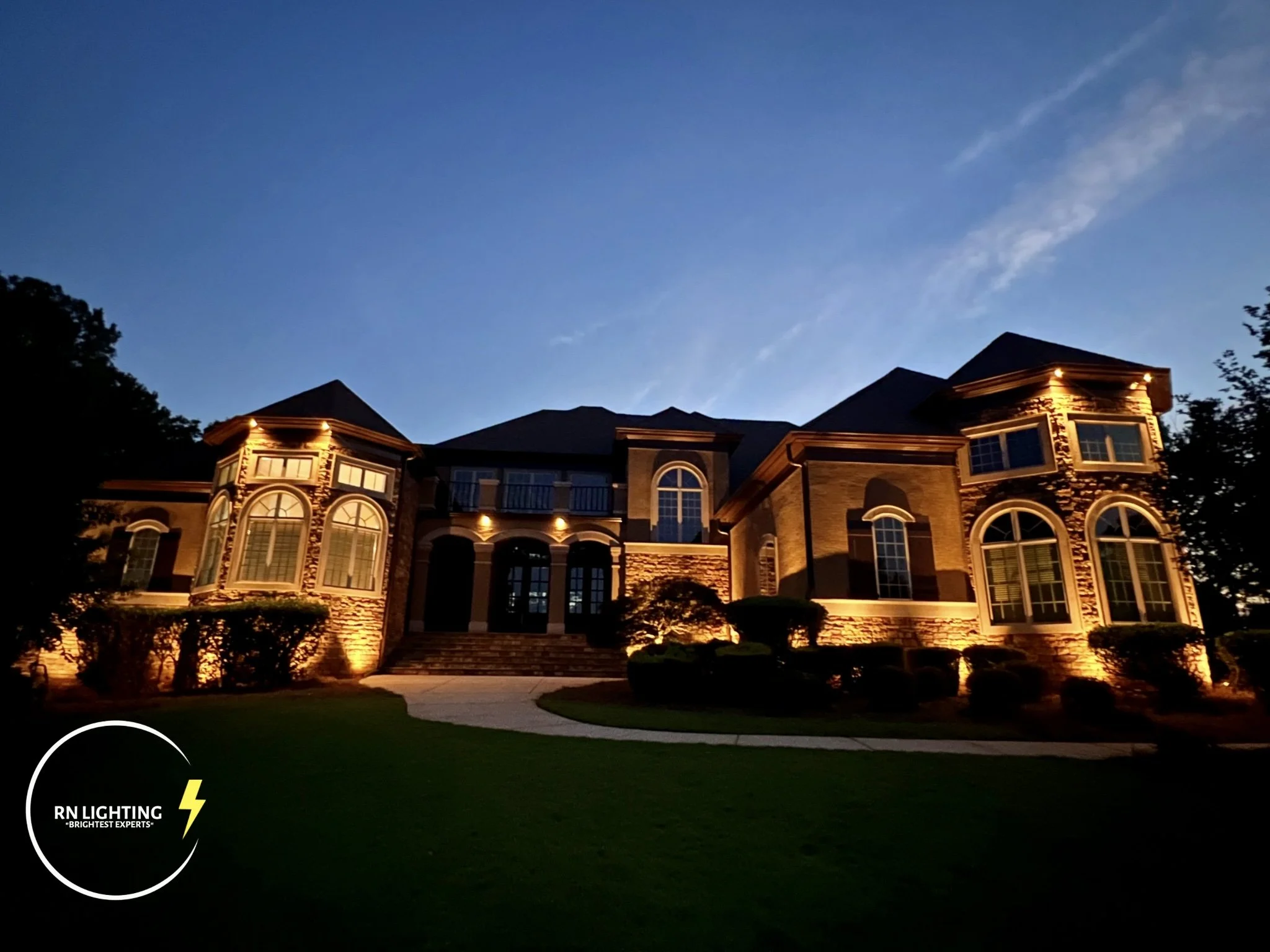A large house with exterior lighting at dusk, showing multiple arched and rectangular windows, a staircase leading to the front door, and a well-manicured lawn with bushes and trees.