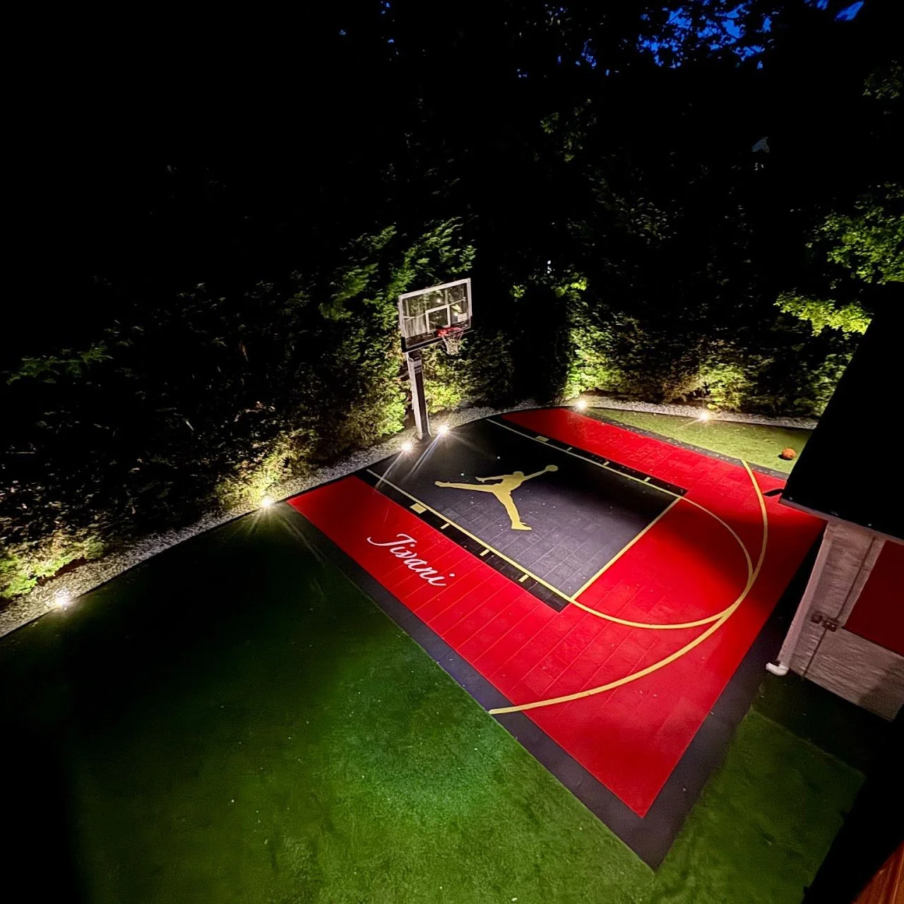 Nighttime view of a basketball court with a black and red surface, illuminated by outdoor lights. The court has a Golden Jumpman logo and the word 'Taori' written on it. A basketball hoop is visible, surrounded by trees.