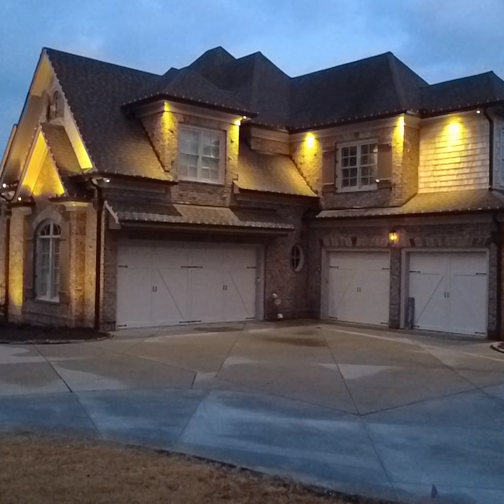 A two-story brick house with a dark shingled roof, illuminated by exterior lights during dusk. The house features multiple garage doors on the ground level and several windows, with the driveway and front yard visible.