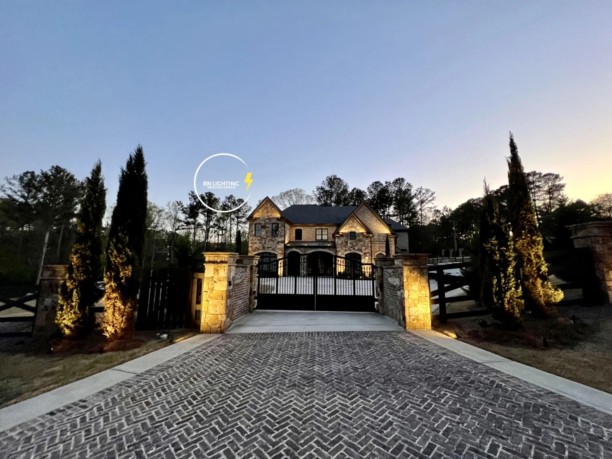 A luxurious house with a stone exterior, large windows, and a gated entrance illuminated by exterior lighting during dusk, with tall trees surrounding the property.