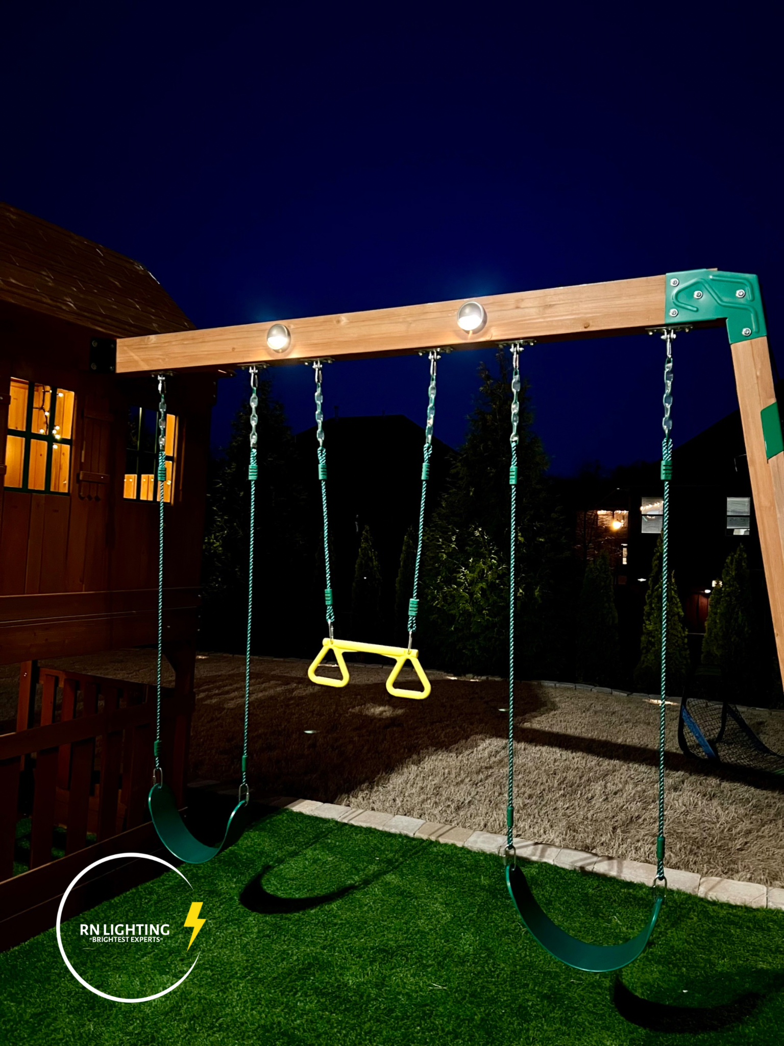 A nighttime view of a backyard playground with swings, a yellow trapeze, and a black swing, illuminated by outdoor lighting, with a house, trees, and other homes in the background.